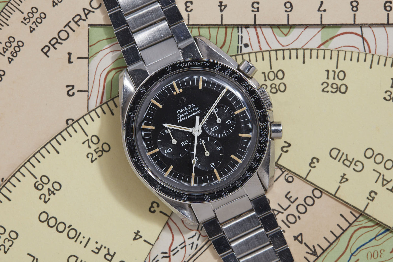 An Omega Speedmaster Professional by Omega, featuring a stainless steel case, black dial, and tachymeter bezel, is displayed atop a vintage map with protractor—evoking NASA’s legacy in the Space Race era.