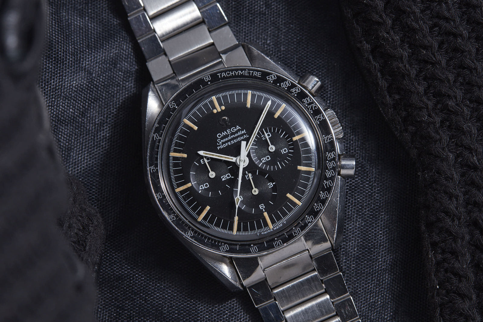 A close-up of the Omega Speedmaster Professional by Omega, featuring a metal band, black dial with three subdials and vintage markers—an iconic watch associated with NASA—shown on a dark textured fabric background.