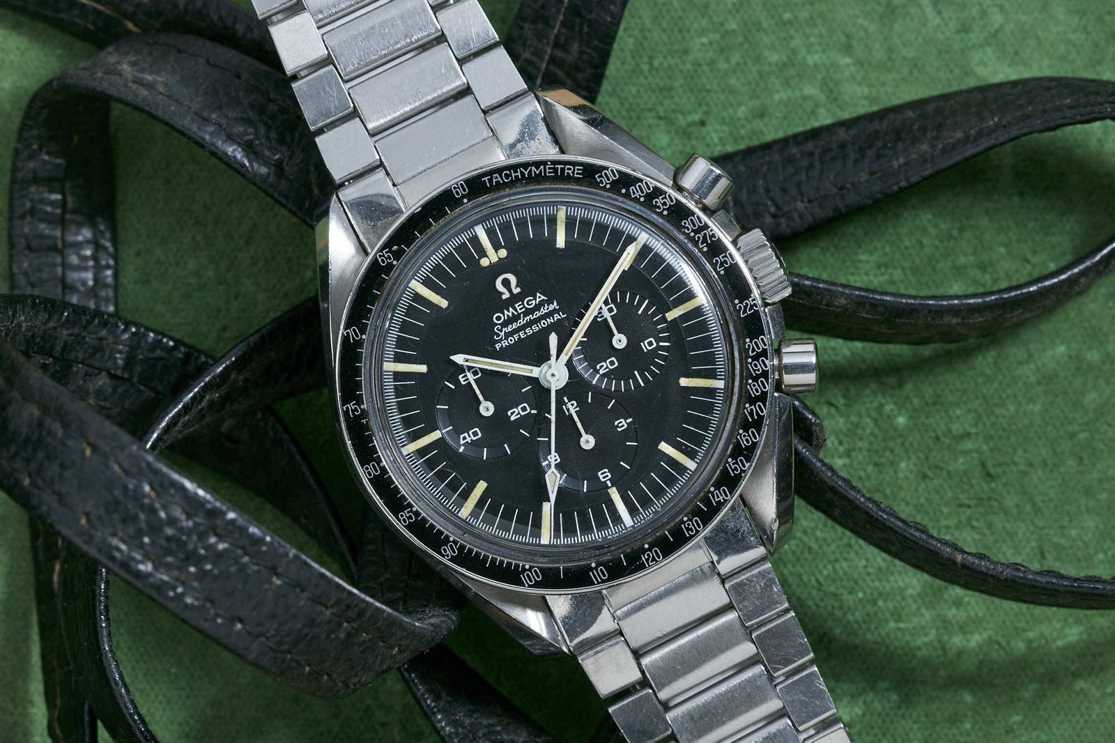 An Omega Speedmaster Professional by Omega, featuring a black dial, tachymeter bezel, and metal bracelet, is displayed atop coiled black leather straps on a textured green background.
