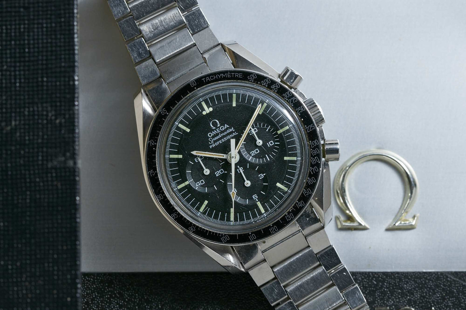 A close-up of the Omega Speedmaster Professional by Omega, featuring a stainless steel band, black dial with three subdials, and the iconic Caliber 321 movement beside a metallic Omega logo on a textured surface.