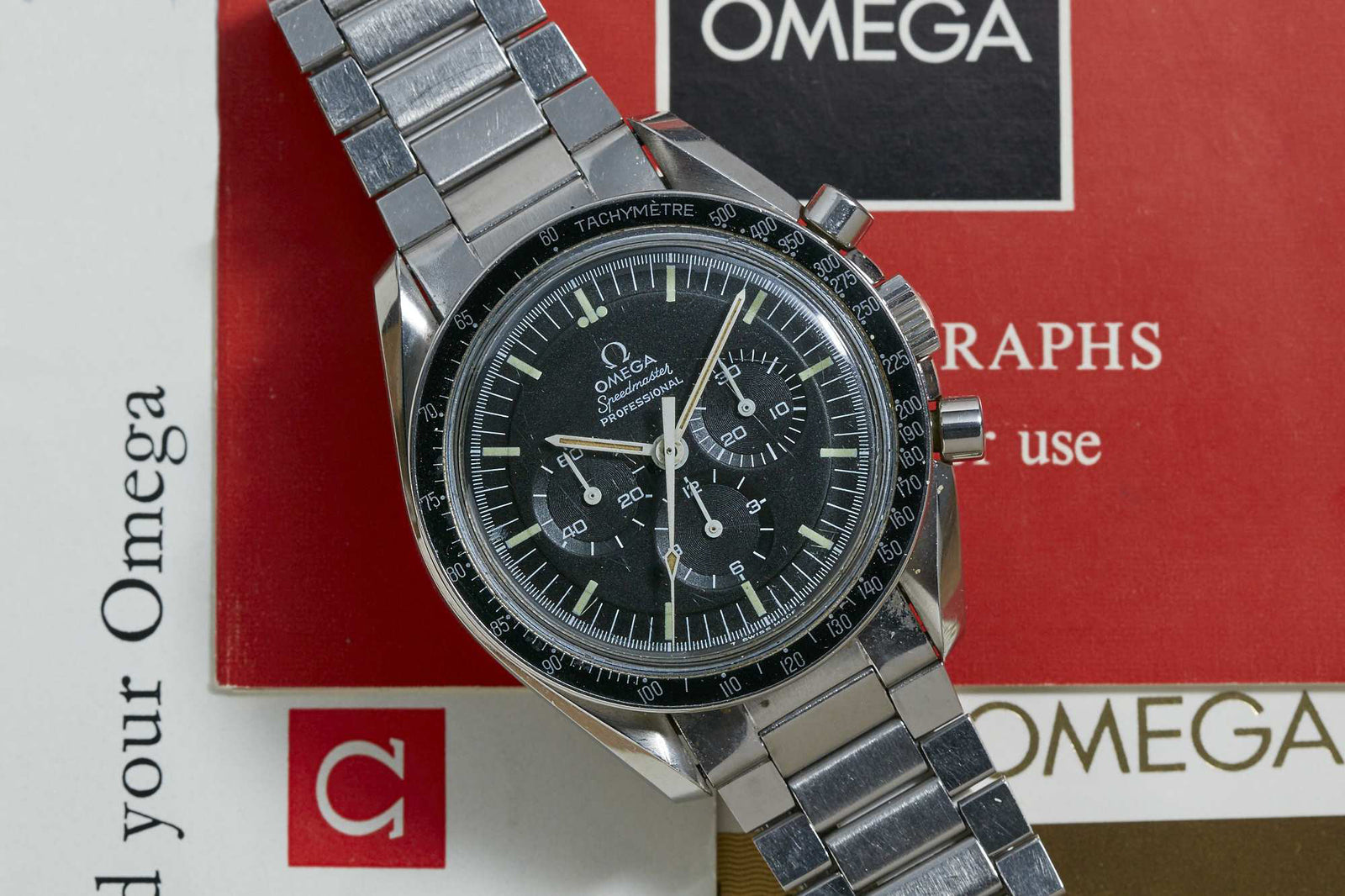 An Omega Speedmaster Professional chronograph with a black dial and tachymeter bezel, shown atop red and white Omega manuals and documents.