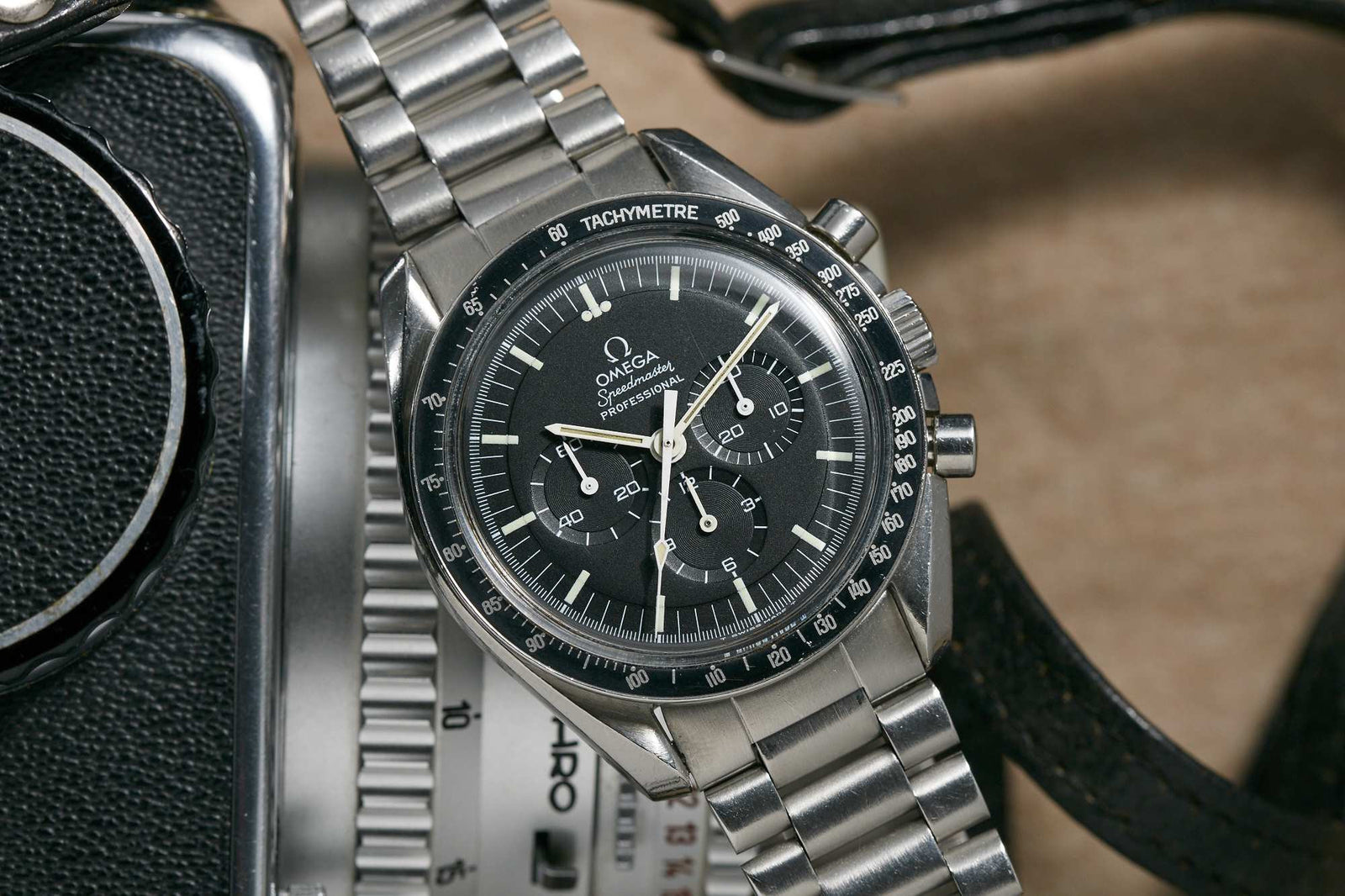 A close-up of the Omega Speedmaster Professional by Omega, featuring a black dial and stainless steel bracelet, displayed beside a vintage camera and leather strap on a beige surface.