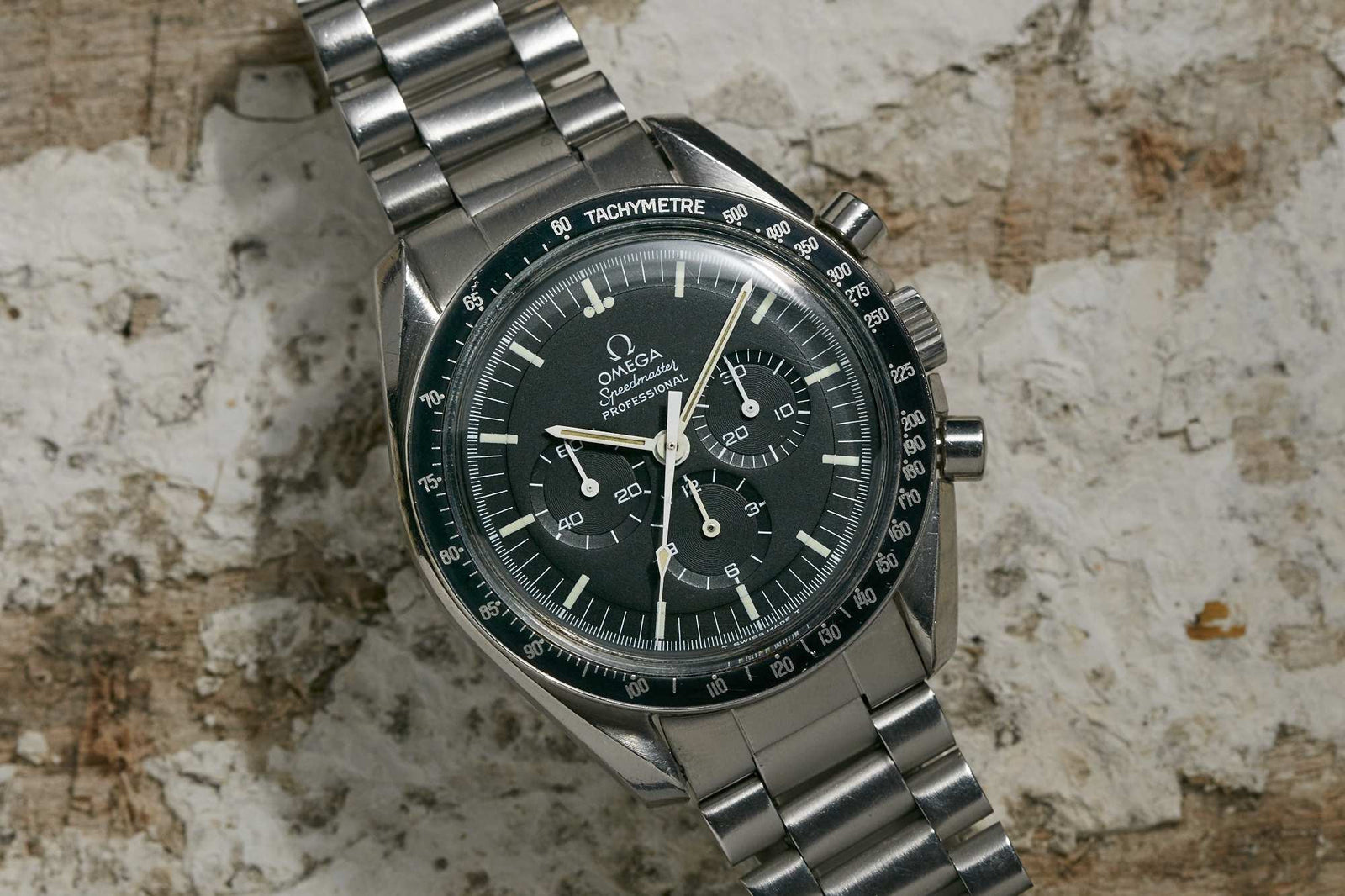 An Omega Speedmaster Professional by Omega, featuring a black dial, three subdials, and a tachymeter bezel, displayed on a textured light surface.