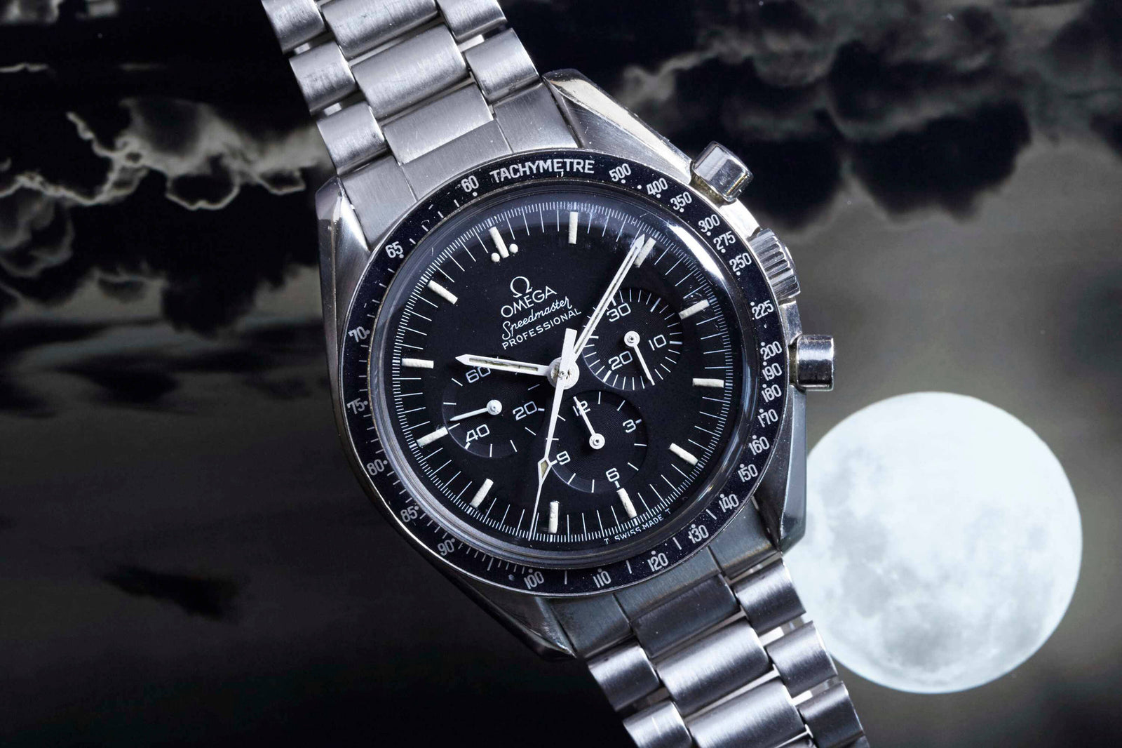 An Omega Speedmaster Professional by Omega with a black dial and tachymeter bezel is shown against a cloudy night sky illuminated by a bright full moon.
