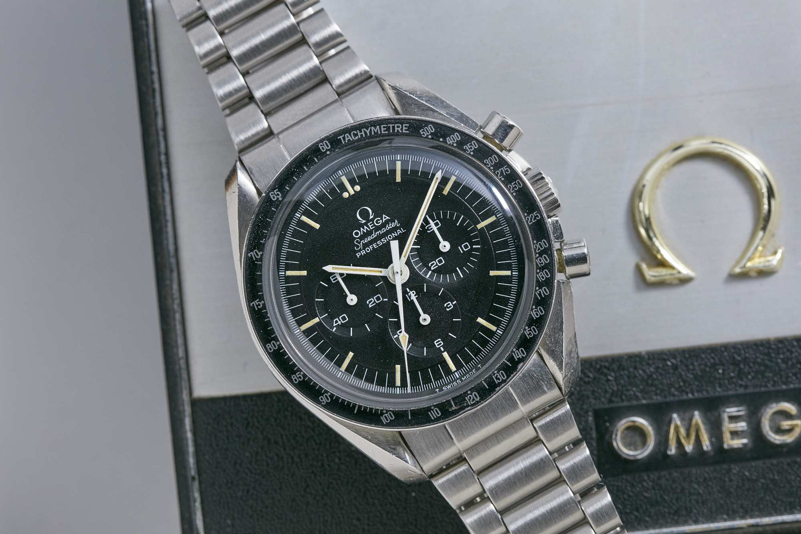 An Omega Speedmaster Professional stainless steel wristwatch with a black tachymeter bezel and chronograph dials rests on a textured box displaying the gold Omega logo.