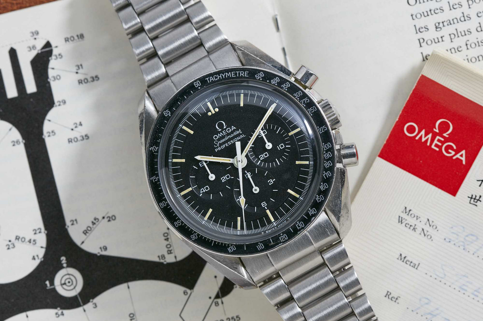 An Omega Speedmaster Professional wristwatch by Omega, featuring a black dial and stainless steel band, is displayed resting on open documents, including a chart and an official Omega certificate.