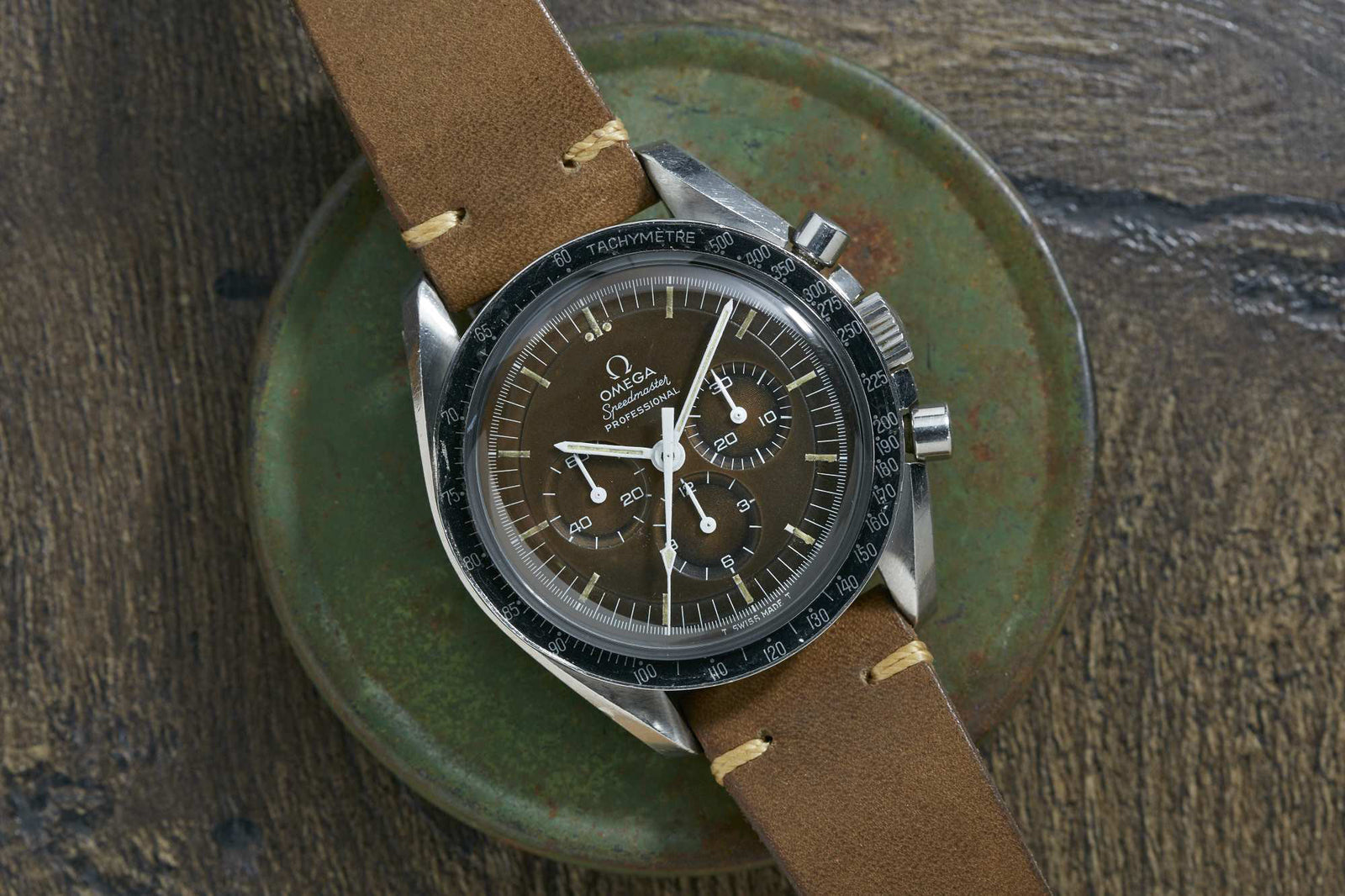 An Omega Speedmaster Professional Tropical wristwatch by Omega, featuring a brown dial, white markers, and a tan leather strap, is displayed on a round green surface atop a textured brown background.
