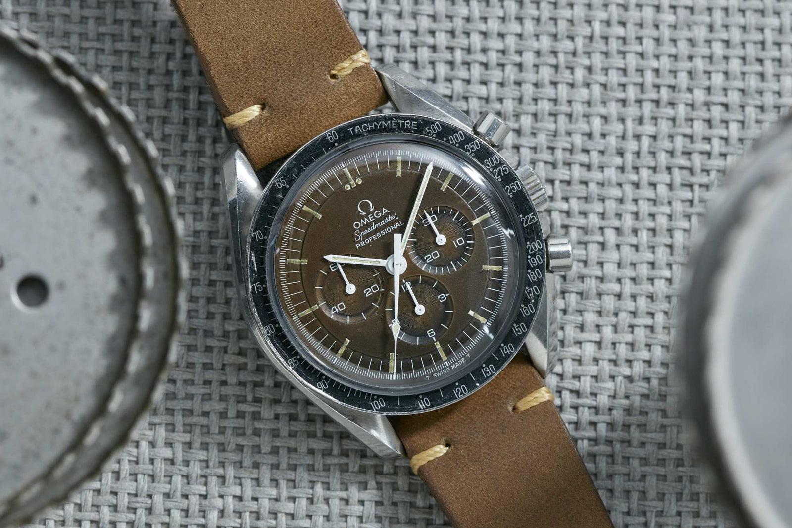 An Omega Speedmaster Professional Tropical by Omega, featuring a brown dial, black tachymeter bezel, and tan leather strap, rests on a textured gray woven surface.