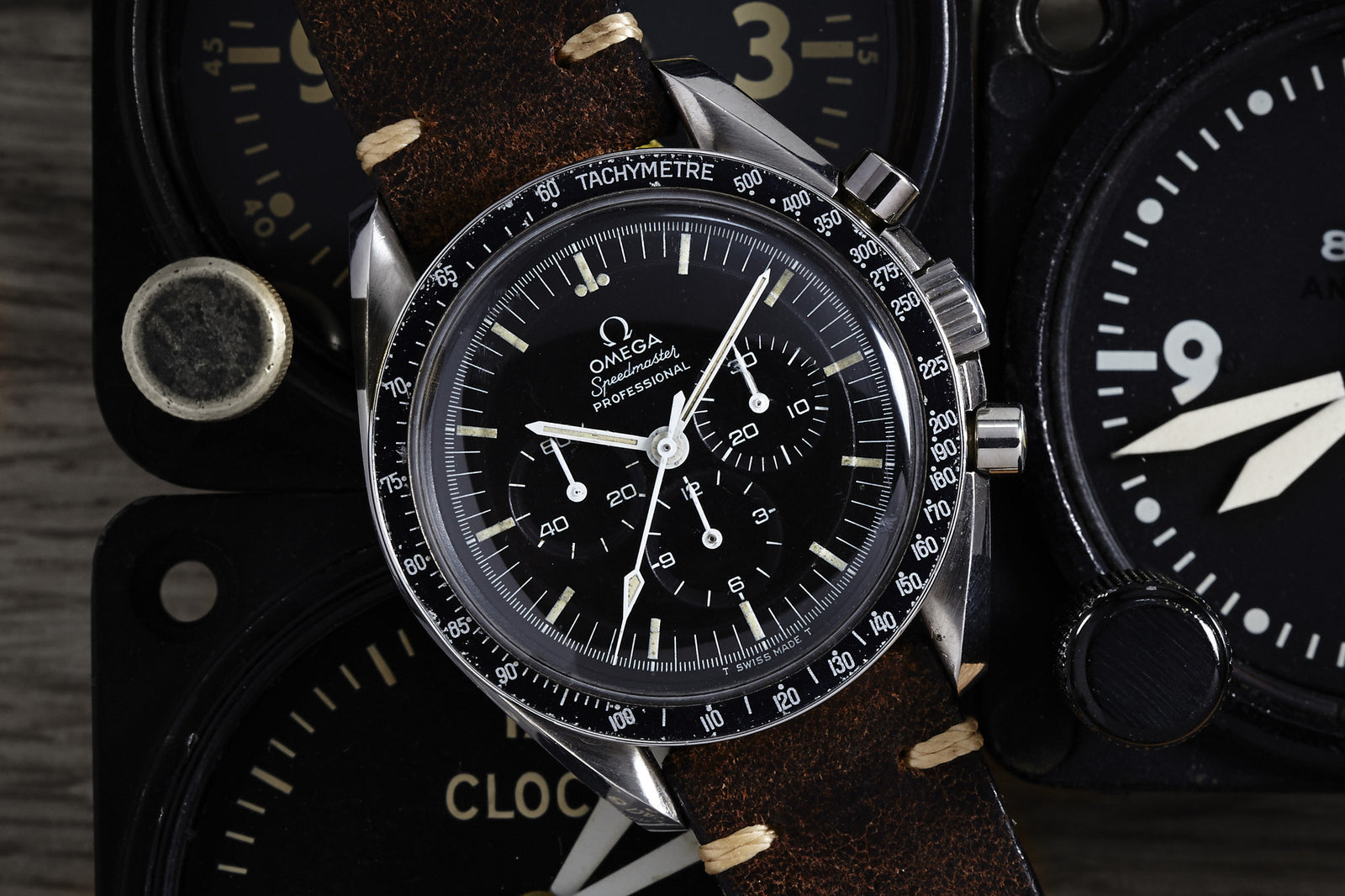 A close-up of the Omega Speedmaster Professional - 1969 by Omega, featuring a black dial and brown leather strap, resting on vintage instrument dials. The watch hands are set to 10:10.