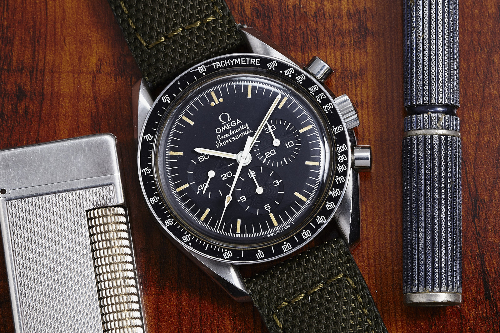 A close-up of the Omega Speedmaster Professional - 1983 by Omega, featuring a green fabric strap, set on a wooden surface next to a silver lighter and a blue-striped pen.