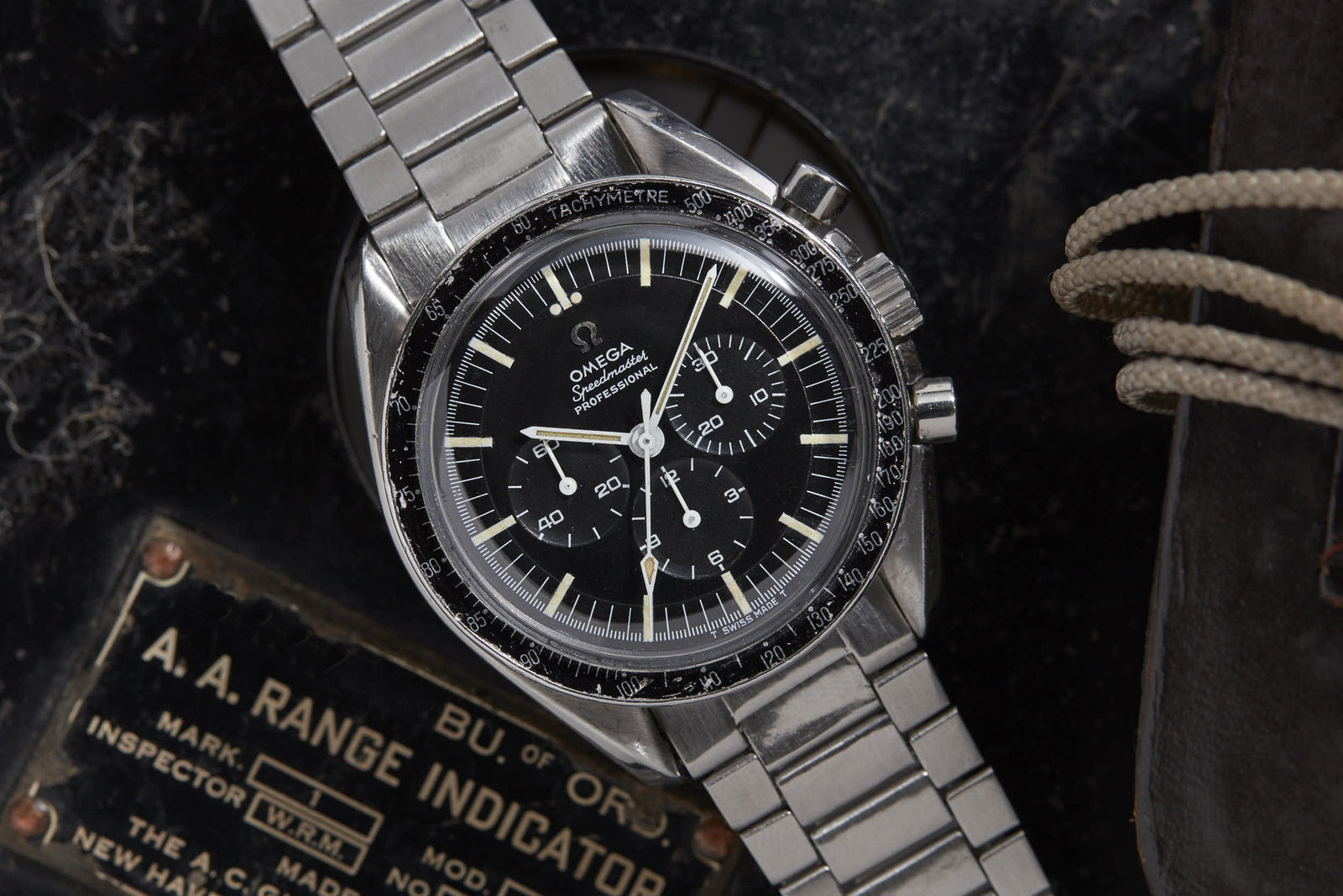 An Omega Speedmaster by Omega with a stainless steel band sits on a black surface beside an A. A. Range Indicator plate—referencing the Apollo Guidance Computer—and a brown leather strap with rope detail.
