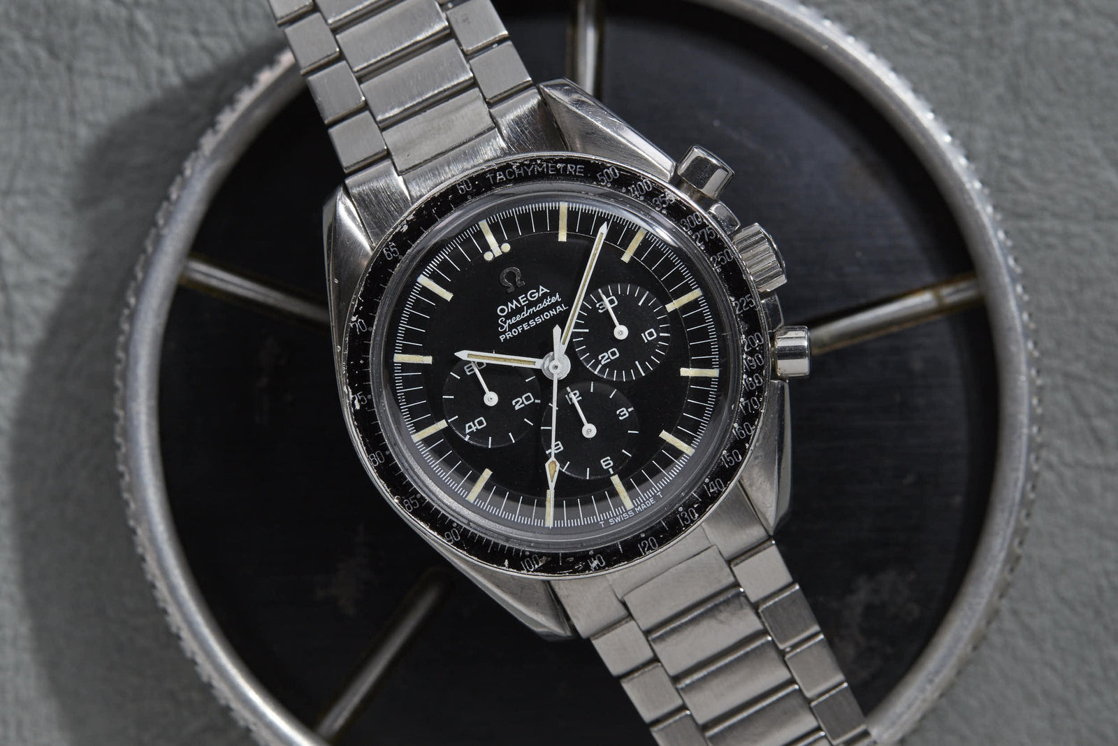 A close-up of the Omega Speedmaster by Omega, featuring a black dial, silver hands, chronograph sub-dials, and a stainless steel band—an iconic wristwatch displayed on a round metallic surface.