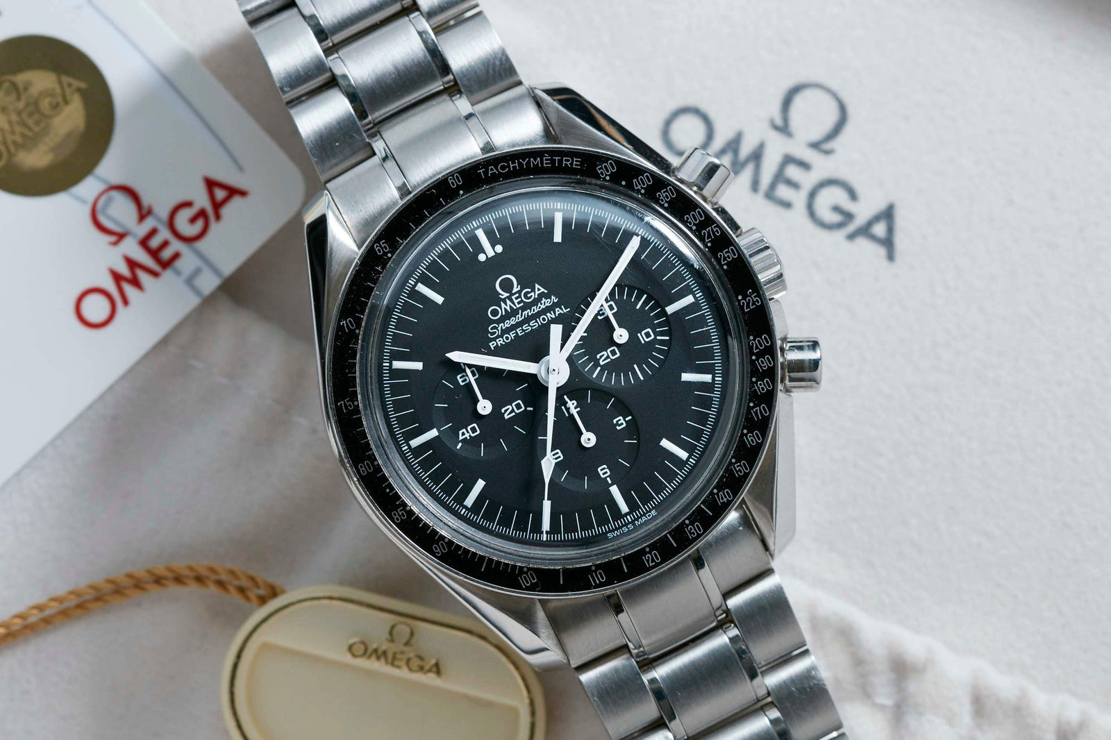A close-up of the Omega Speedmaster Professional "Hesalite Sandwich" by Omega, featuring a black dial and stainless steel band on a soft cloth alongside Omega tags and packaging.
