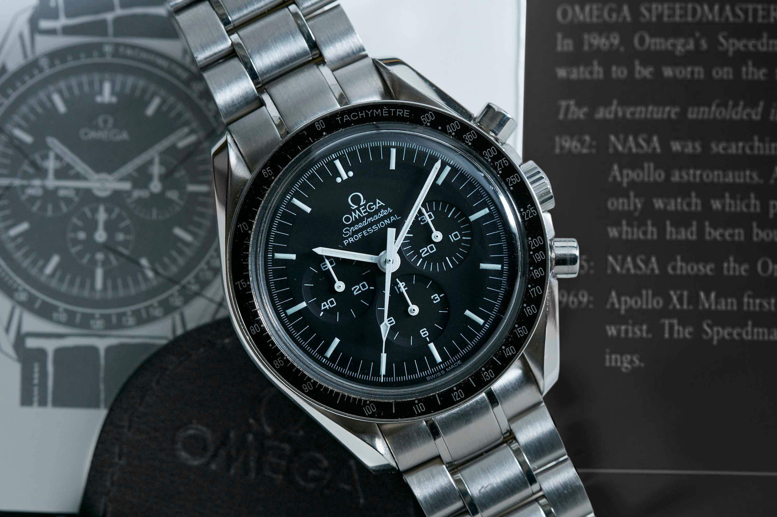 A close-up of the Omega Speedmaster Professional "Hesalite Sandwich" by Omega, featuring a stainless steel band, black dial, and chronograph functions, displayed with its booklet and case.