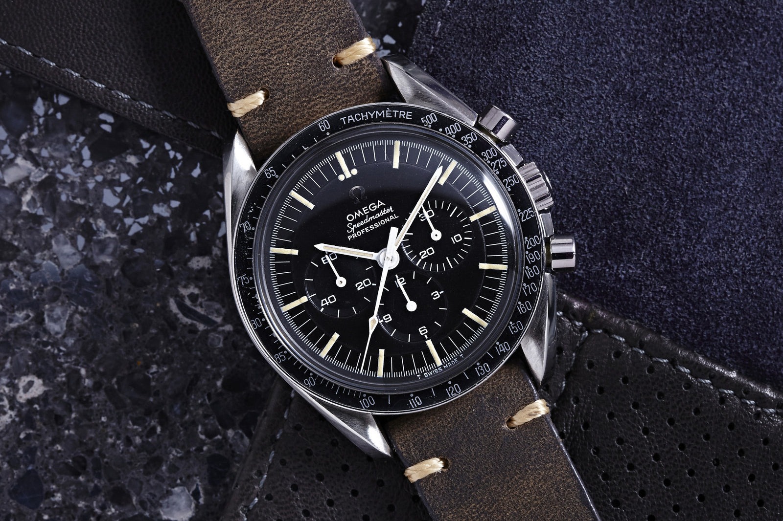 A close-up of the Omega Speedmaster Professional Transitional - 1968 by Omega, featuring a black dial, white hands and markers, brown leather strap, and tachymeter bezel, resting on a textured dark gray surface.