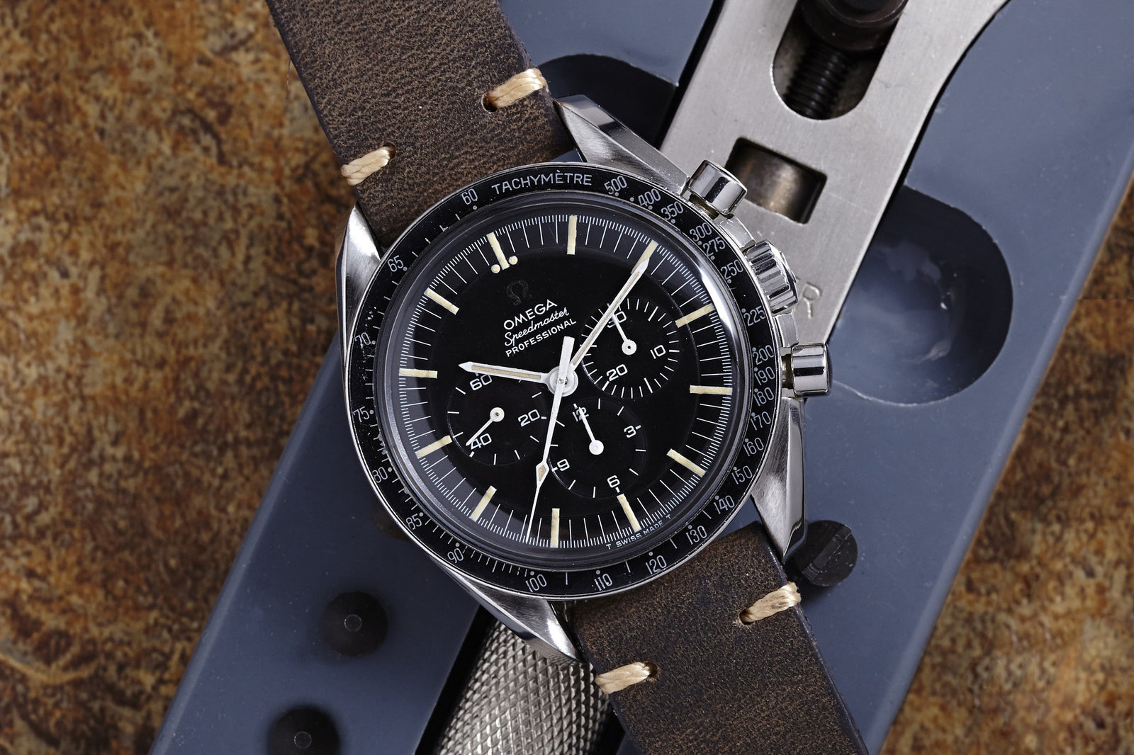 A close-up of the Omega Speedmaster Professional Transitional - 1968, showcasing its black dial and brown leather strap on a textured metallic surface with a warm brownish background.