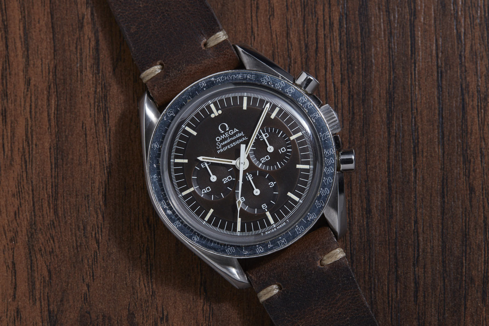 An Omega Speedmaster Professional wristwatch by Omega, featuring a black dial with three subdials and a tachymeter bezel, is shown on a brown leather strap resting on a wooden surface. Famously associated with the Moon Landing.