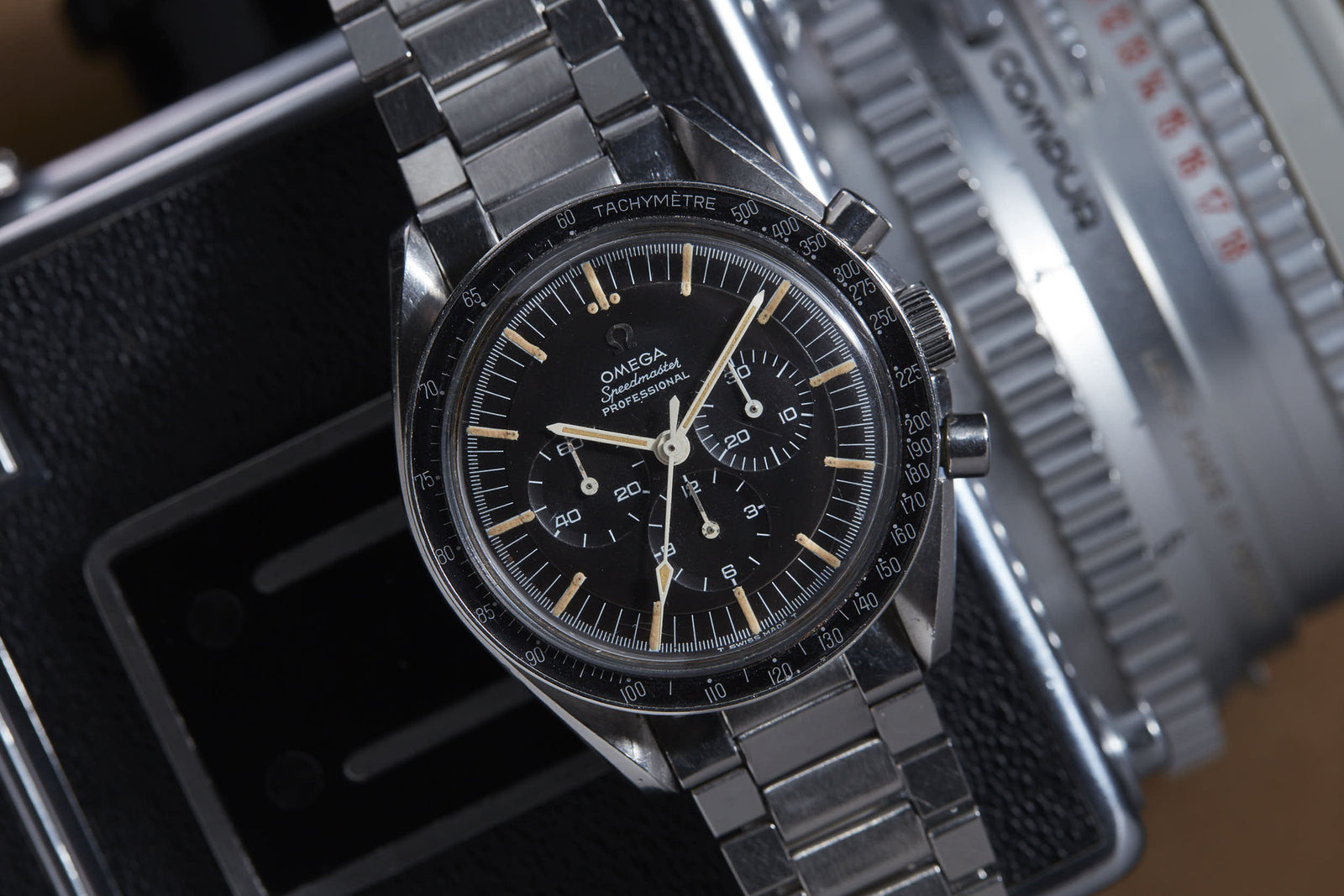 A close-up of the Omega Speedmaster Professional by Omega, featuring a stainless steel band, black dial, and chronograph, famously worn during the Apollo missions, resting on a vintage camera.