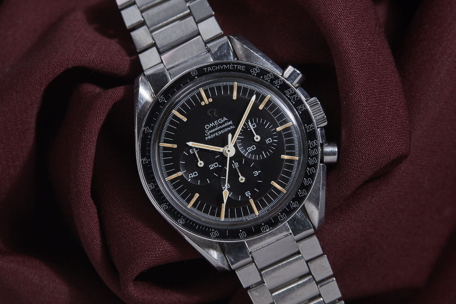 A close-up of the Omega Speedmaster Professional by Omega shows its black dial, chronograph subdials, and silver metal strap, resting on rich burgundy fabric—a model renowned for its Apollo mission legacy.