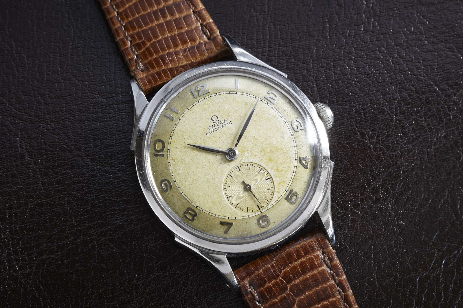 The Omega Steel Bumper Wind by Omega features a worn cream dial with Arabic numerals, a small seconds subdial at 6 o’clock, and a brown textured leather strap, displayed on a dark leather surface for classic appeal.