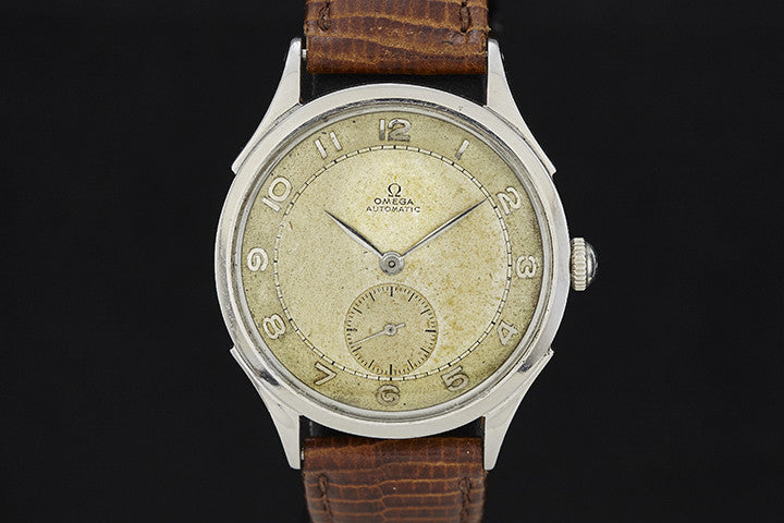 The Omega Steel Bumper Wind by Omega features a worn beige dial with silver numerals and a small seconds subdial at 6 o’clock, set in a silver case with a brown textured leather strap against a deep black background.