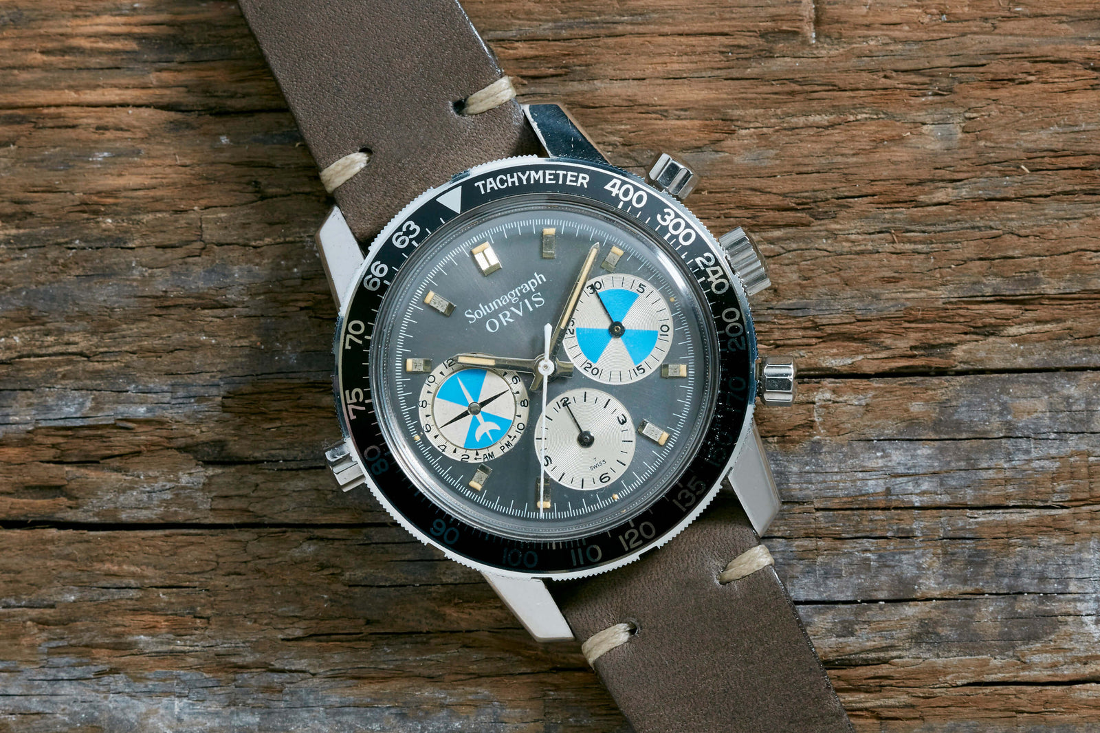 A Heuer Solunagraph wristwatch with a brown leather strap and black tachymeter bezel rests on wood. The blue, white, and black subdials feature tide indication, with "Südhangarung ORLY 1979" on the dial. Brand: Heuer.