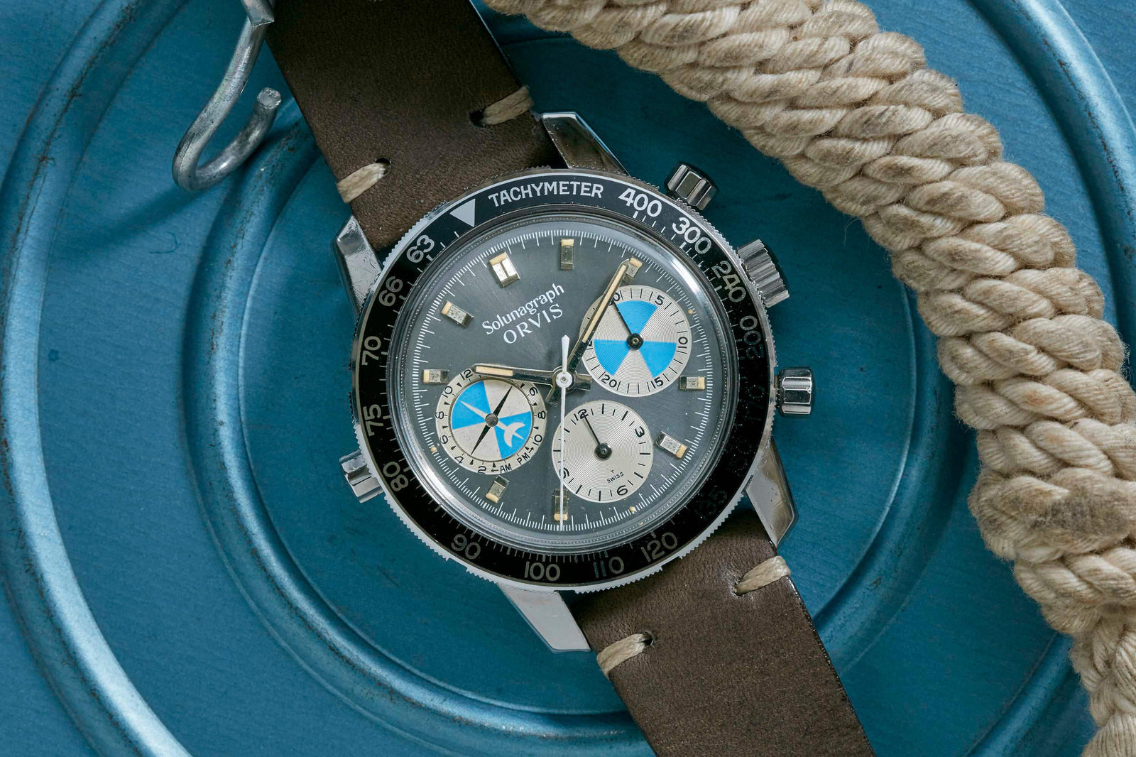 A close-up of the Heuer Solunagraph wristwatch by Heuer, featuring a brown leather strap, dark bezel, and blue and white subdials with tide indication. The watch rests on a coiled rope atop a blue circular surface.