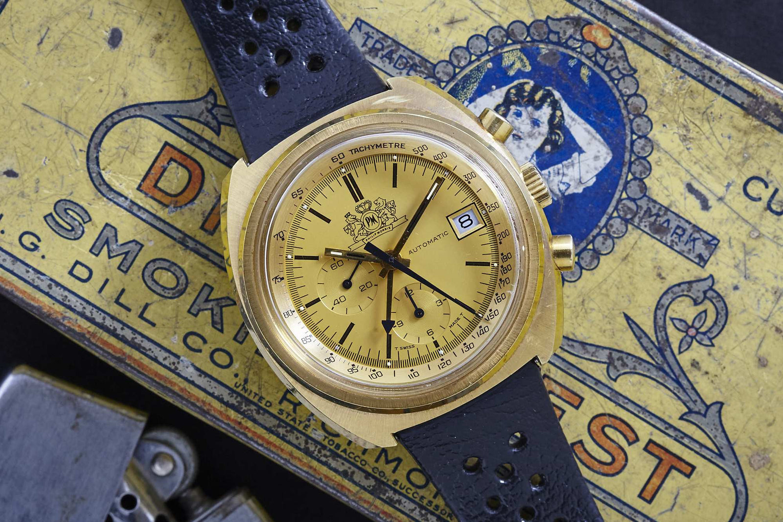 An Omega Philip Morris gold chronograph watch with a black perforated strap sits on a vintage yellow and blue Philip Morris tin, featuring a tachymeter scale, Lemania Caliber 1341, chronograph subdials, and date window at 3 o'clock.