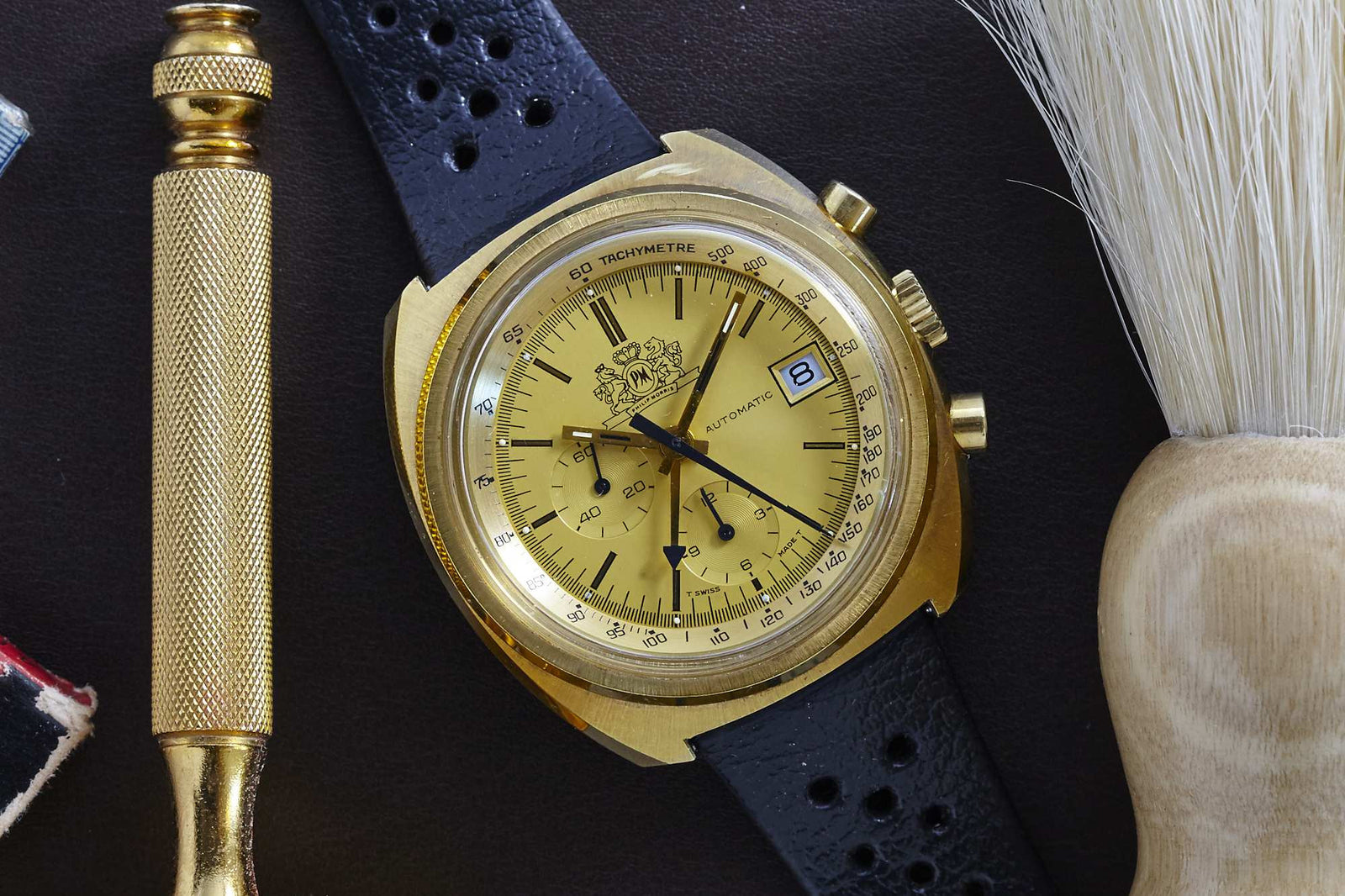 An Omega Philip Morris gold chronograph watch with a yellow dial, black markers, and a black leather strap rests on a dark surface beside a gold tool and a shaving brush with white bristles.
