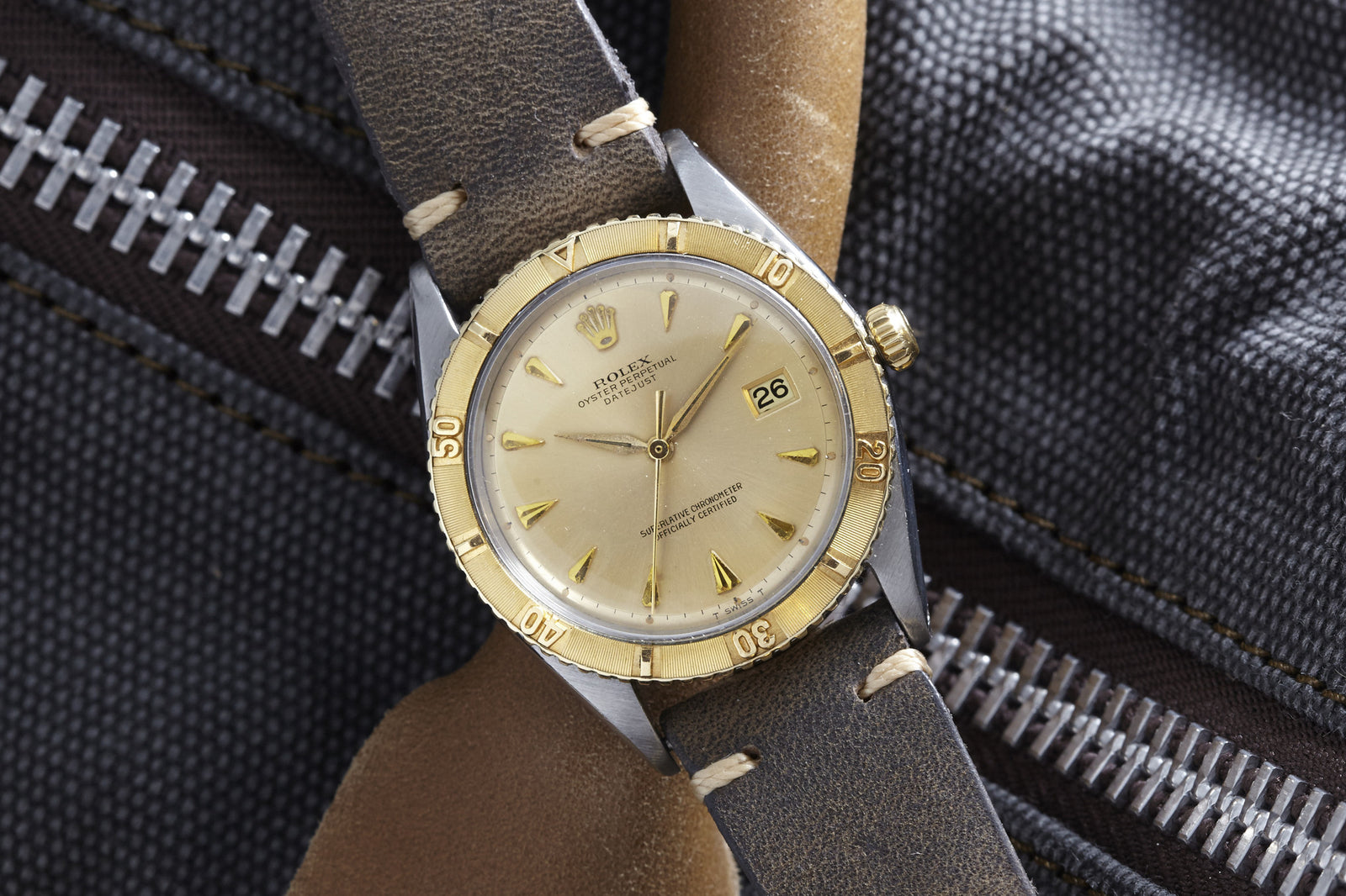 A Rolex Turn-O-Graph wristwatch by Rolex, featuring a gold bezel and tan leather strap, is displayed on dark textured fabric with a visible metal zipper. The date is shown on its classic dial, highlighting the watch's vintage appeal.