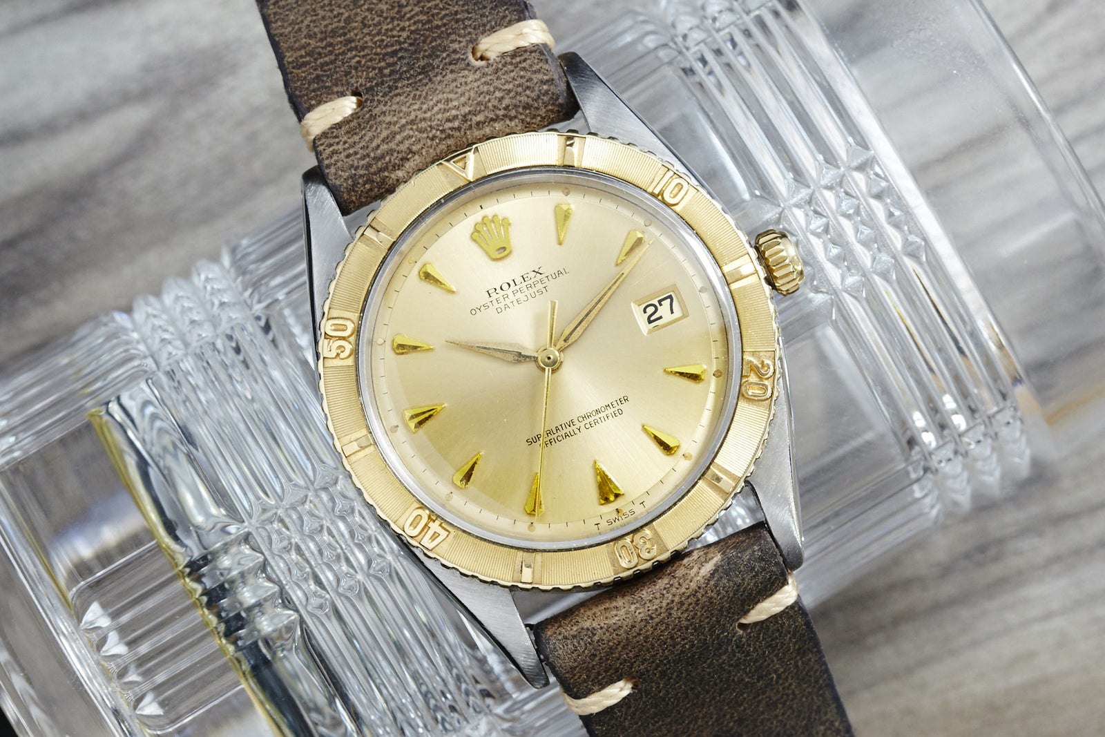 A Rolex Turn-O-Graph wristwatch with a gold bezel, cream dial, gold markers, and brown leather strap rests on textured glass cups, displaying the date at 3 o’clock.