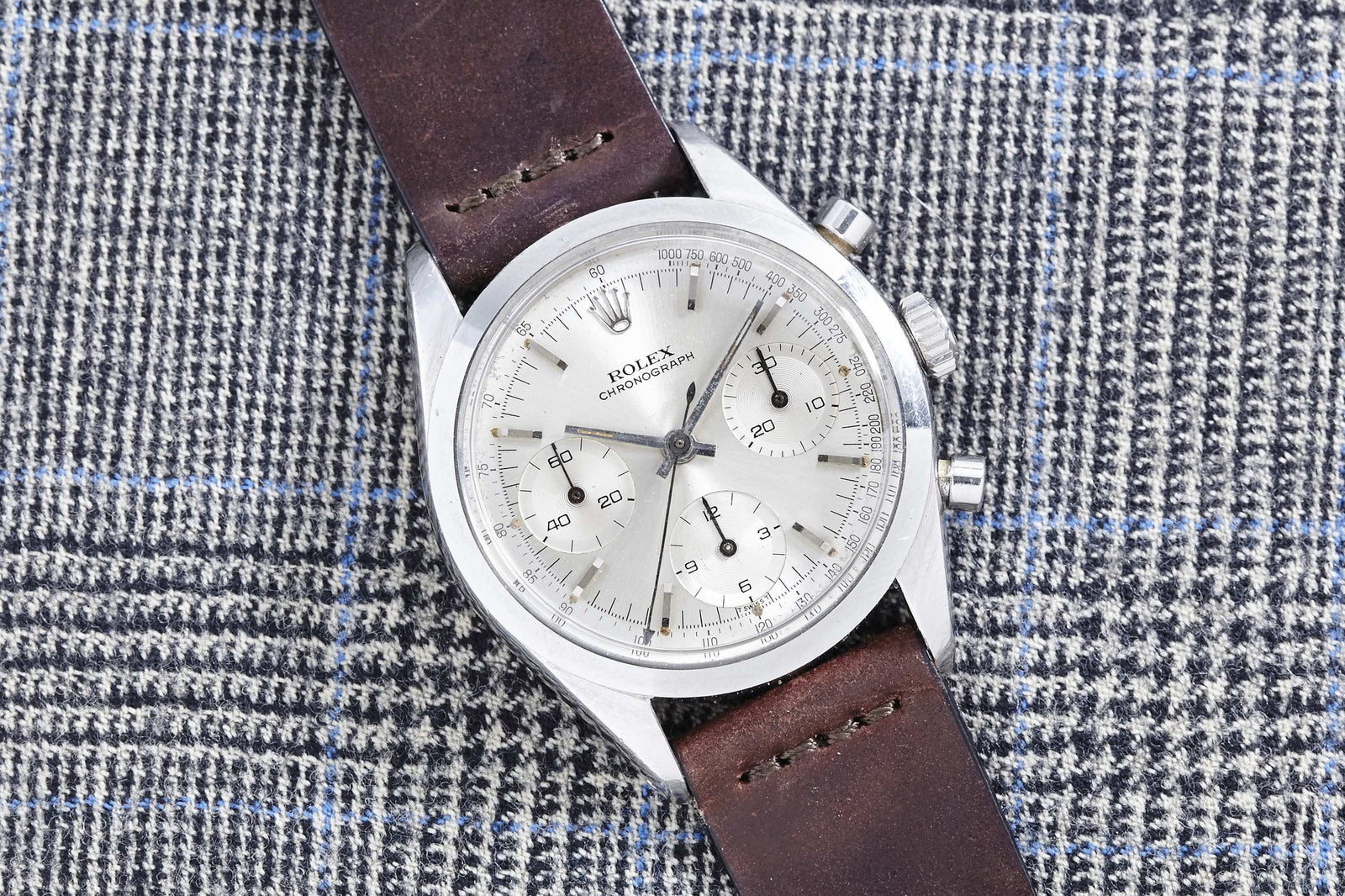 A vintage Rolex Cosmograph 6238 chronograph wristwatch with a brown leather strap rests on gray and blue plaid fabric. This classic Rolex features a silver dial, three subdials, and two side pushers. Description coming soon!.