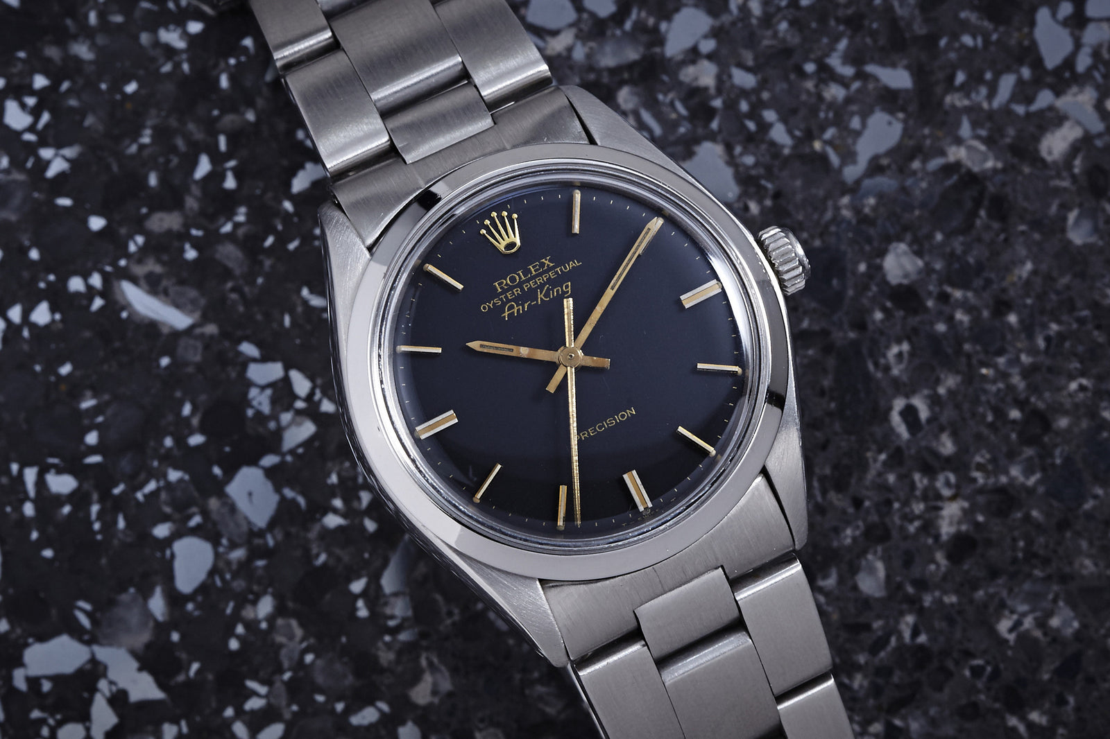 A 1977 Rolex Air King Gilt Dial wristwatch by Rolex, featuring a stainless steel Oyster case and black dial with gold hour markers and hands, displayed on a speckled dark surface.