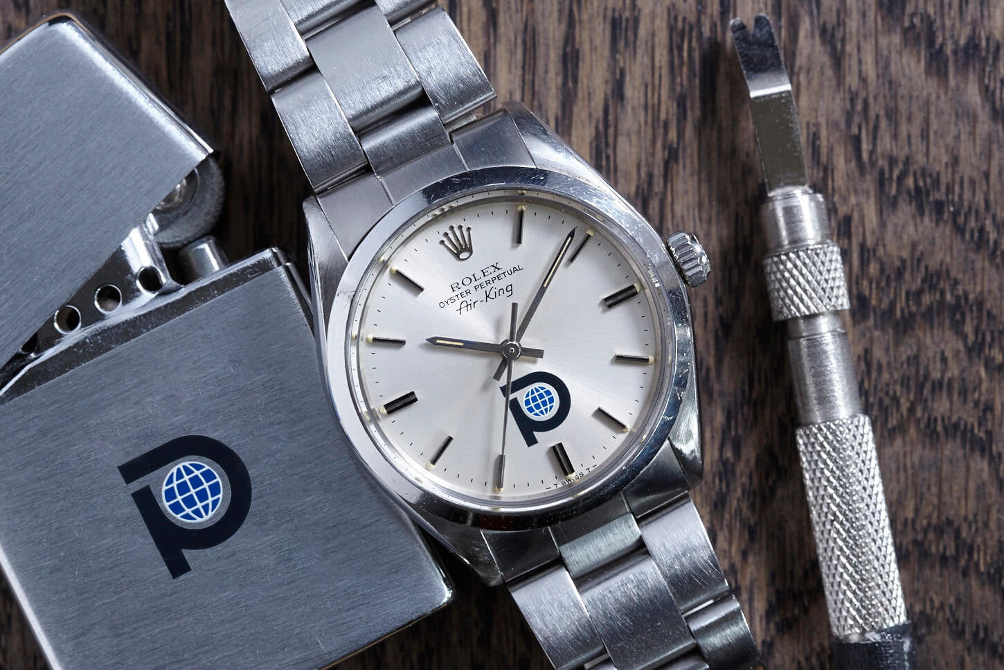 A vintage Rolex Air King Pool Oil by Rolex, with a silver dial and blue-black P logo globe, sits beside a metal lighter and watchmaking tool on wood—a classic Presentation Watch for collectors.