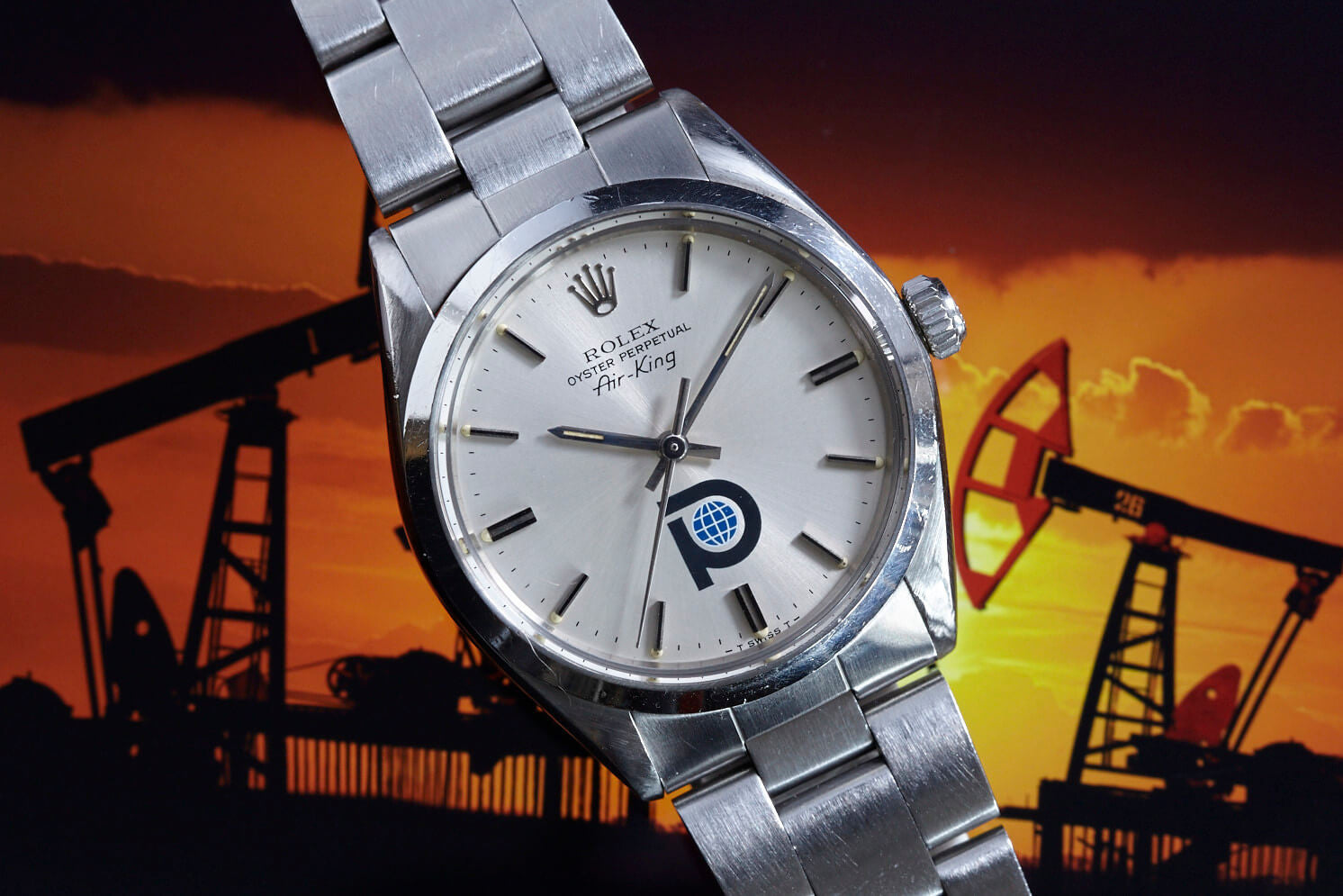 A Rolex Air King Pool Oil wristwatch by Rolex with a white dial, metal band, and a unique blue and black logo at 6 o’clock is shown in close-up against a sunset backdrop with oil pumpjack silhouettes.