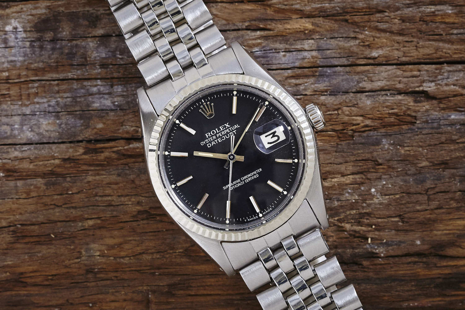 The Rolex Datejust 1601 Black Dial by Rolex features a black dial, silver hour markers, a date window at 3 o’clock, and a metal link bracelet. Its iconic Oyster Perpetual design is showcased on a wooden surface.