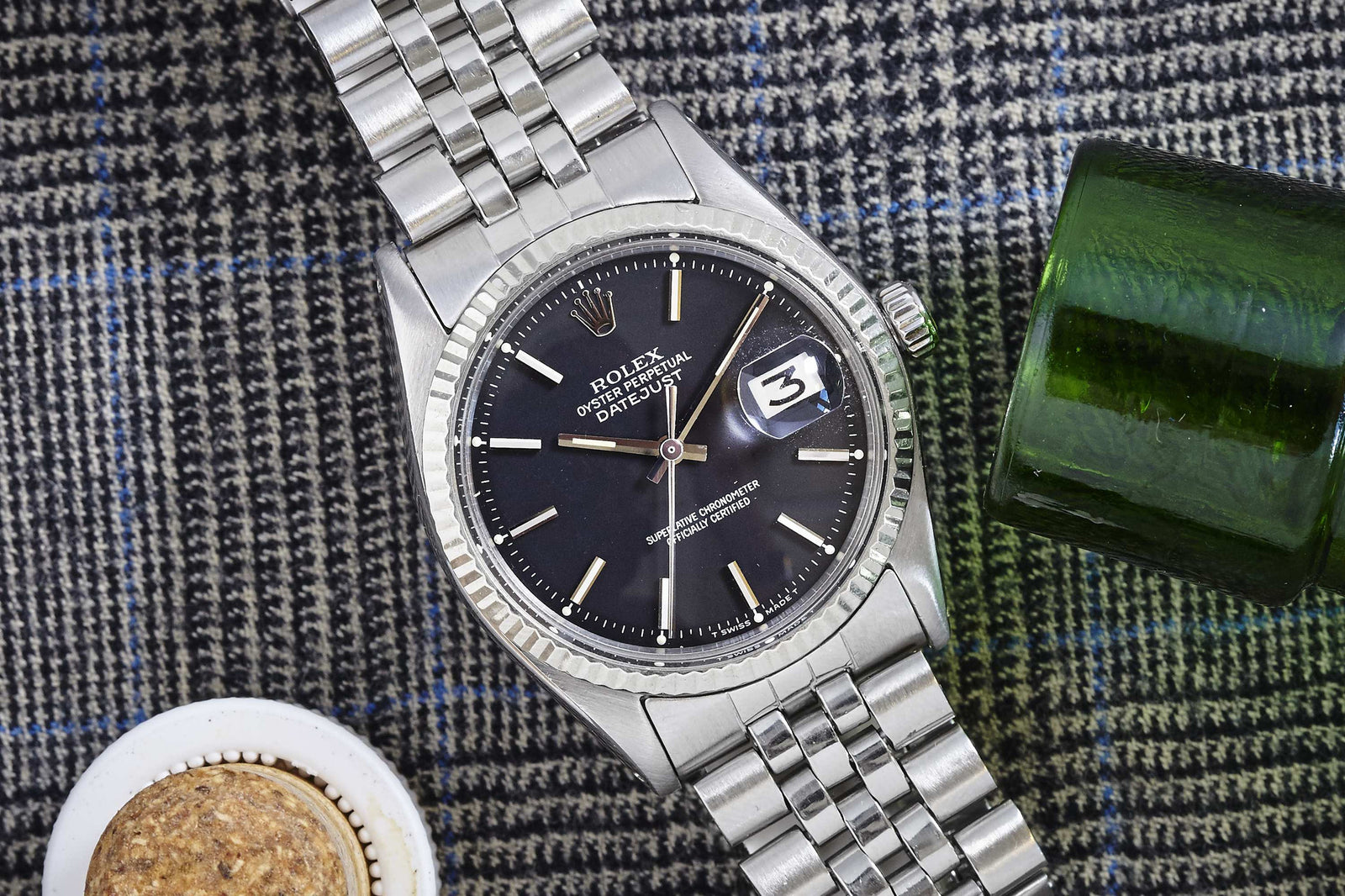 A Rolex Datejust 1601 Black Dial by Rolex sits on gray plaid fabric, with a green glass bottle and a cork-topped circular container beside it.