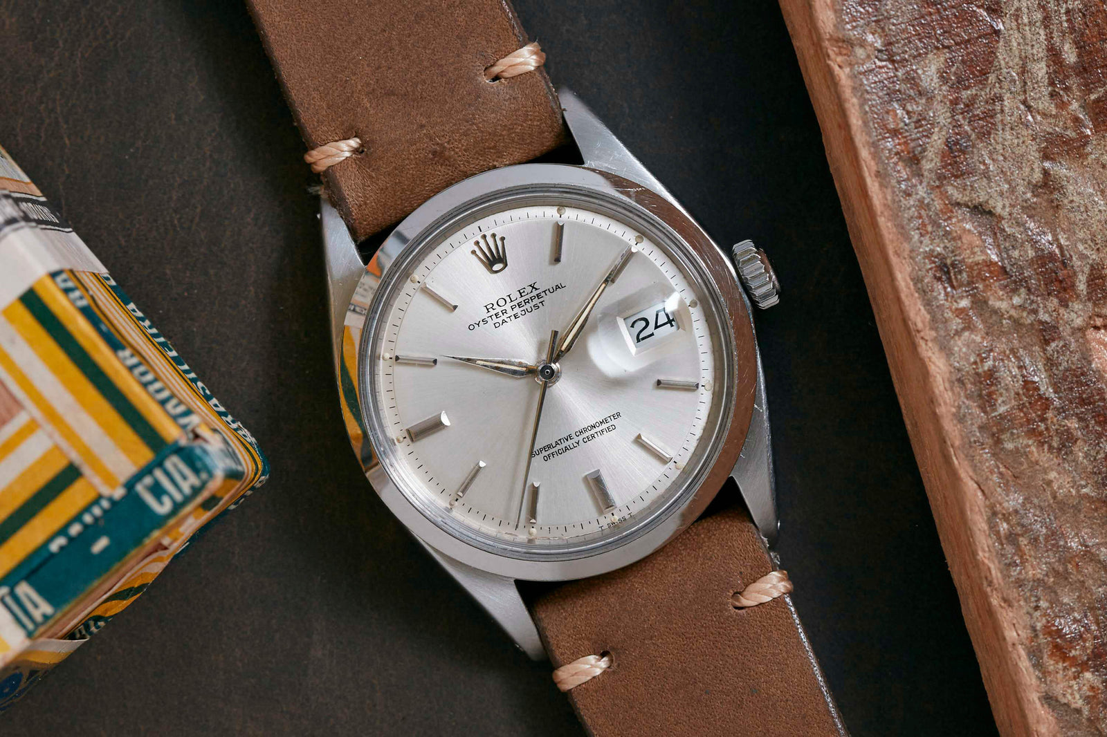 A silver Rolex Datejust by Rolex with a brown leather strap lies on a dark surface, partly framed by a book and a textured rectangle, highlighting its refined design and the date display showing “24”.