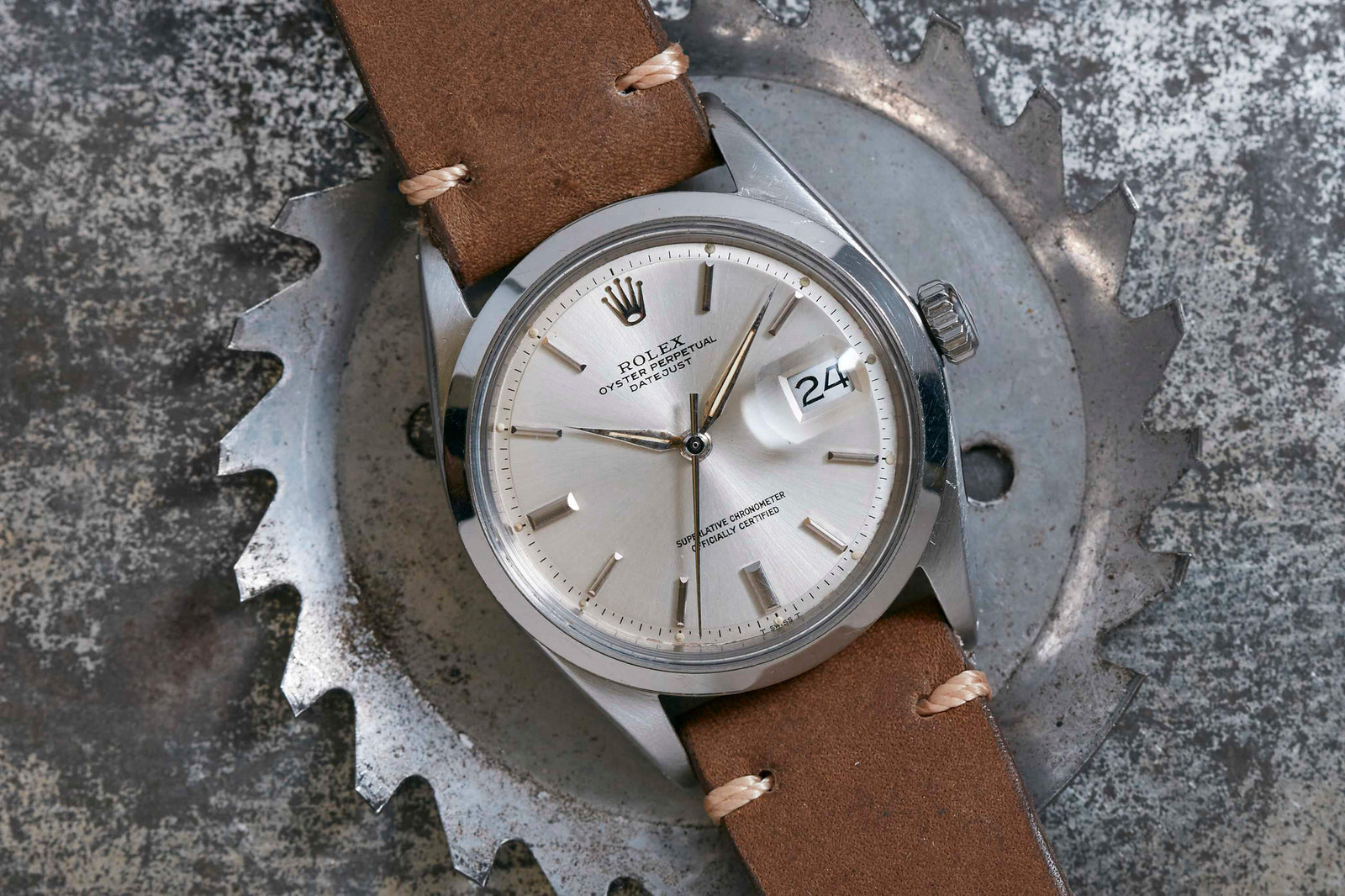 A silver Rolex Datejust by Rolex with a brown leather strap is displayed on a metal gear, showing the date as 24. The background features a textured metallic surface.