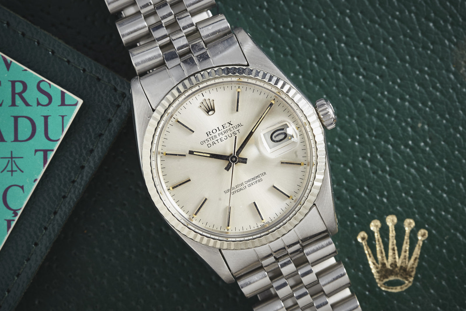 A Rolex Datejust by Rolex with a white dial and jubilee bracelet sits on a textured green surface beside a green wallet and the gold Rolex crown logo, highlighting its timeless style.