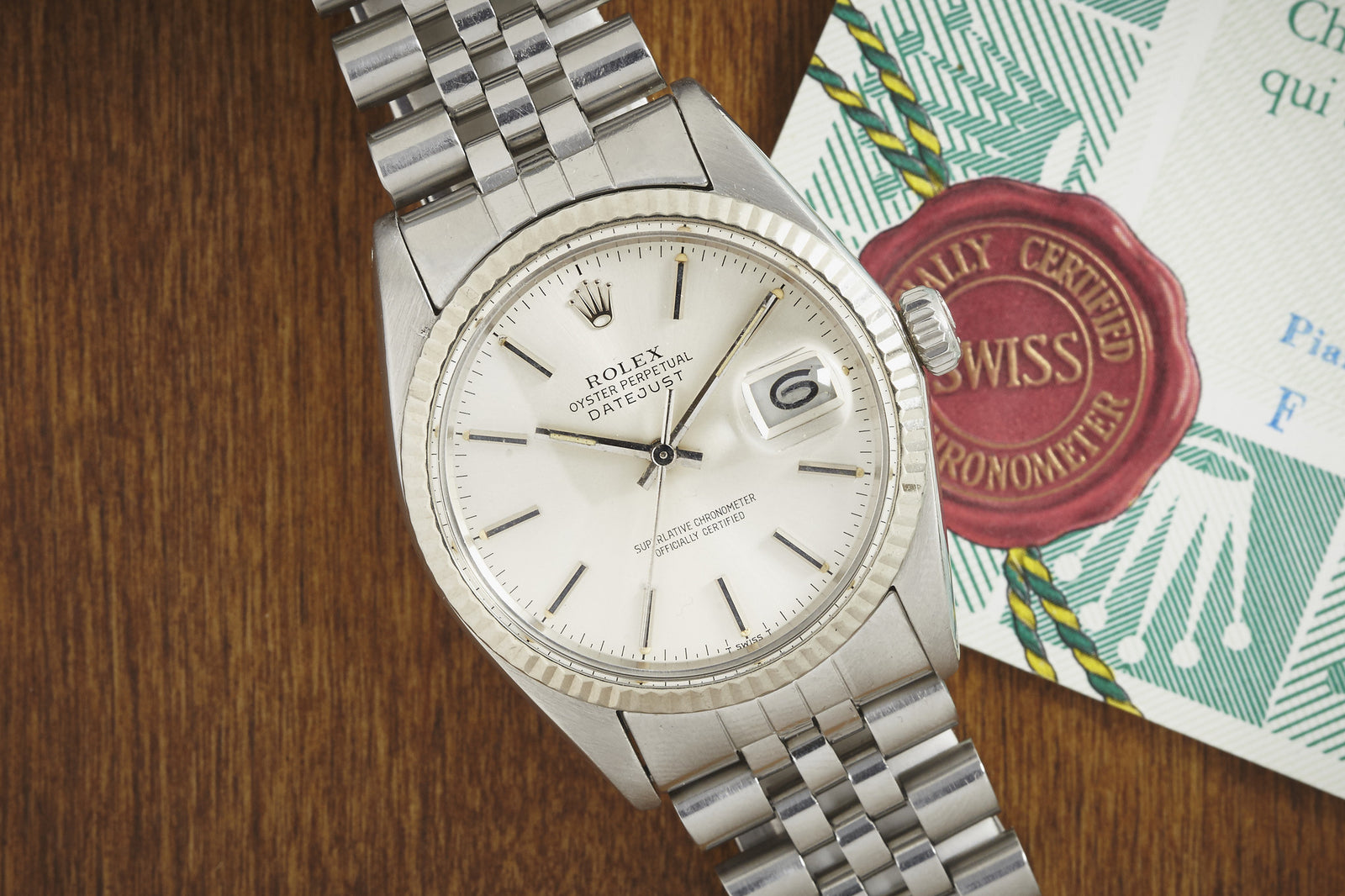 A Rolex Datejust by Rolex, featuring a silver dial, fluted bezel, and metal bracelet, sits next to a partially visible certificate with a red Swiss emblem. The date on the watch reads “6.”.