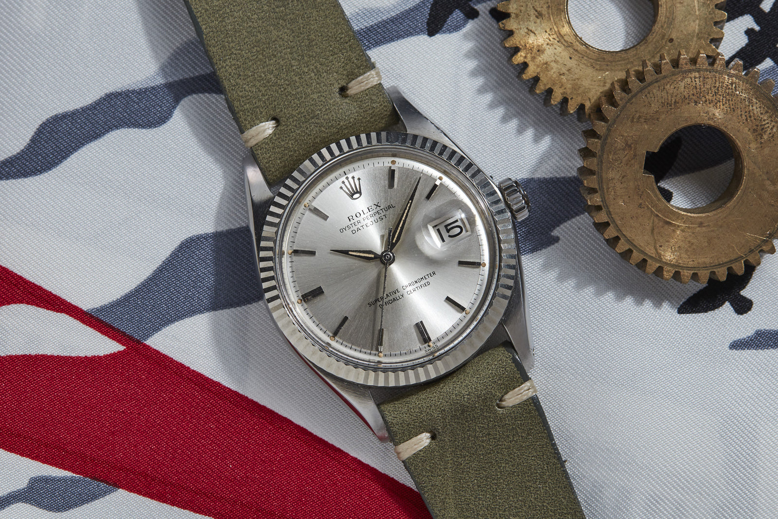 A Rolex Datejust luxury watch by Rolex with a green strap is displayed on patterned fabric next to two metal gears. The silver watch features its signature date function, showing the number “16” on the dial.