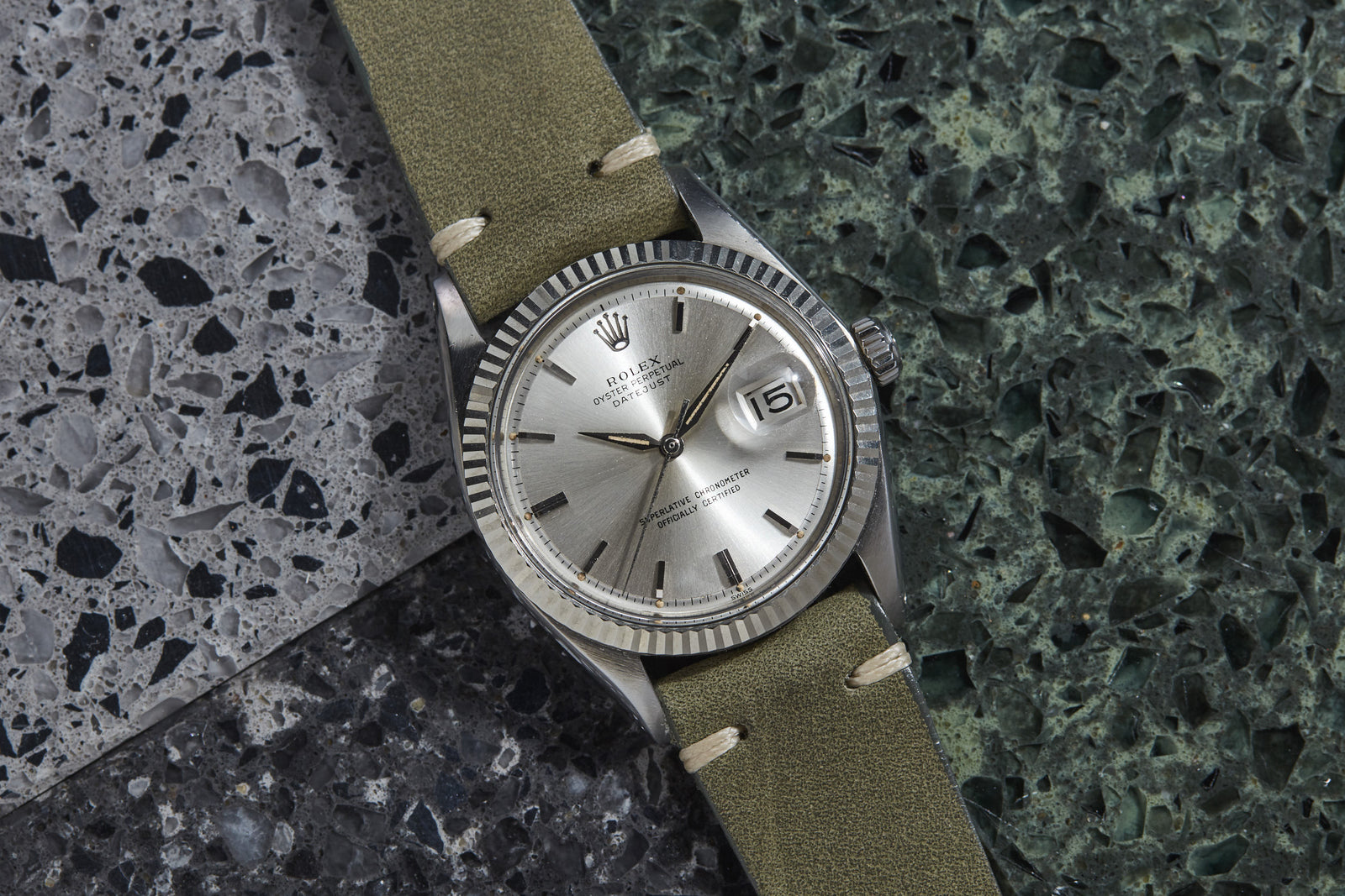 A Rolex Datejust luxury watch with a greenish-brown leather strap sits on speckled gray and green stone tiles. The silver timepiece by Rolex features a date function and classic fluted bezel design.