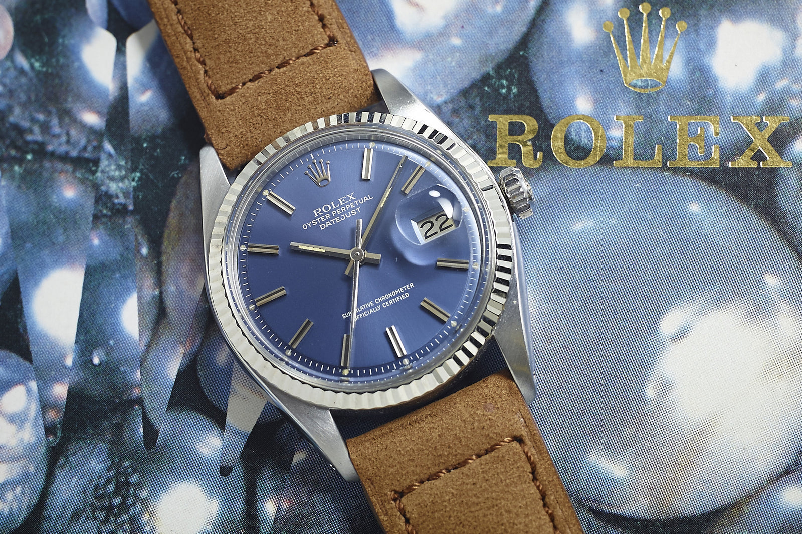 A 1977 Rolex Datejust 1601 with a blue pie pan dial, silver markers, and a brown suede strap is displayed against a gold Rolex logo backdrop, with the date window showing "22".