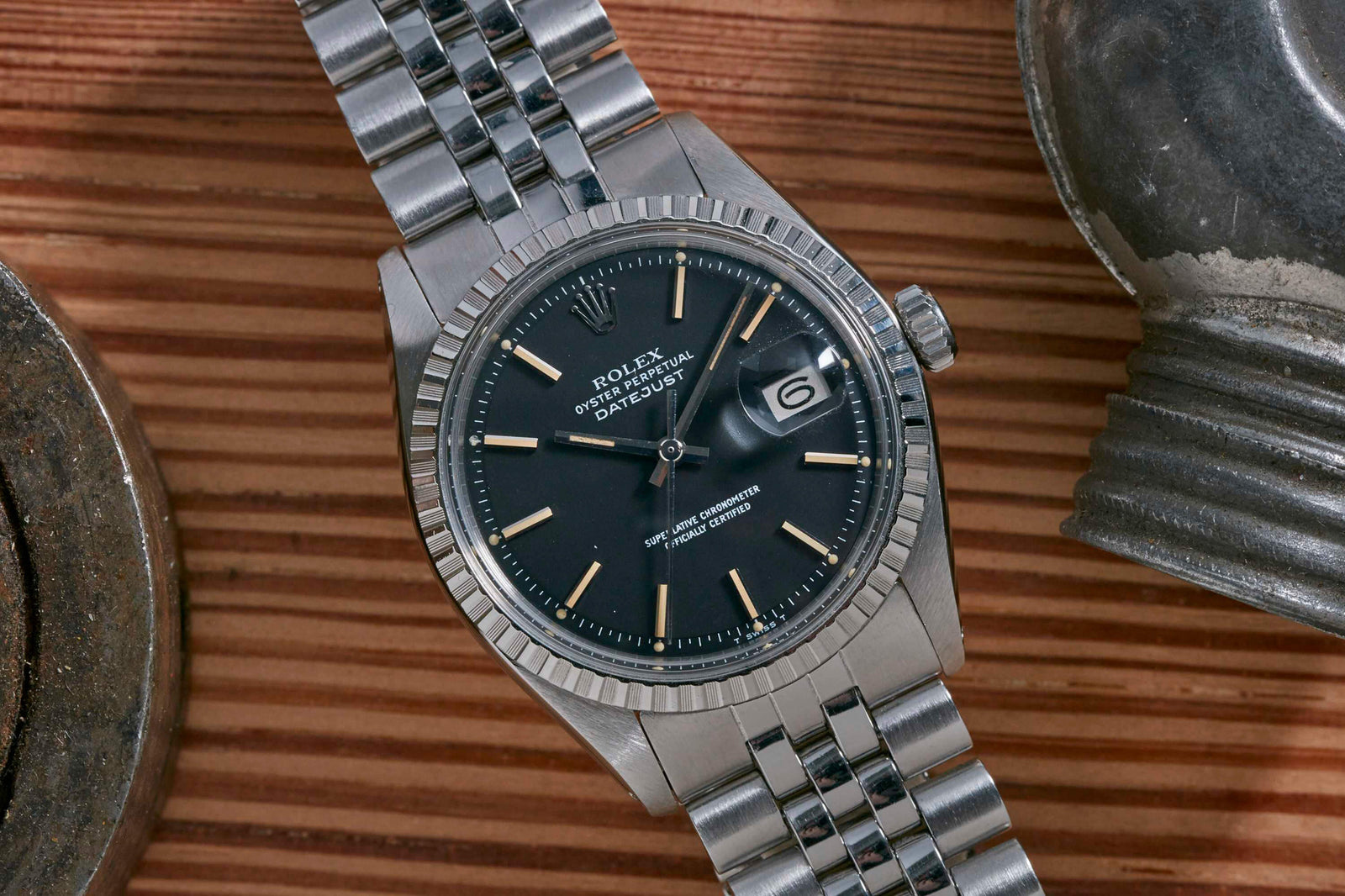 A close-up of the Rolex Datejust by Rolex, featuring a black dial, silver hour markers, and a stainless steel jubilee bracelet, set on wood beside a metallic object—a true icon in luxury timepieces.