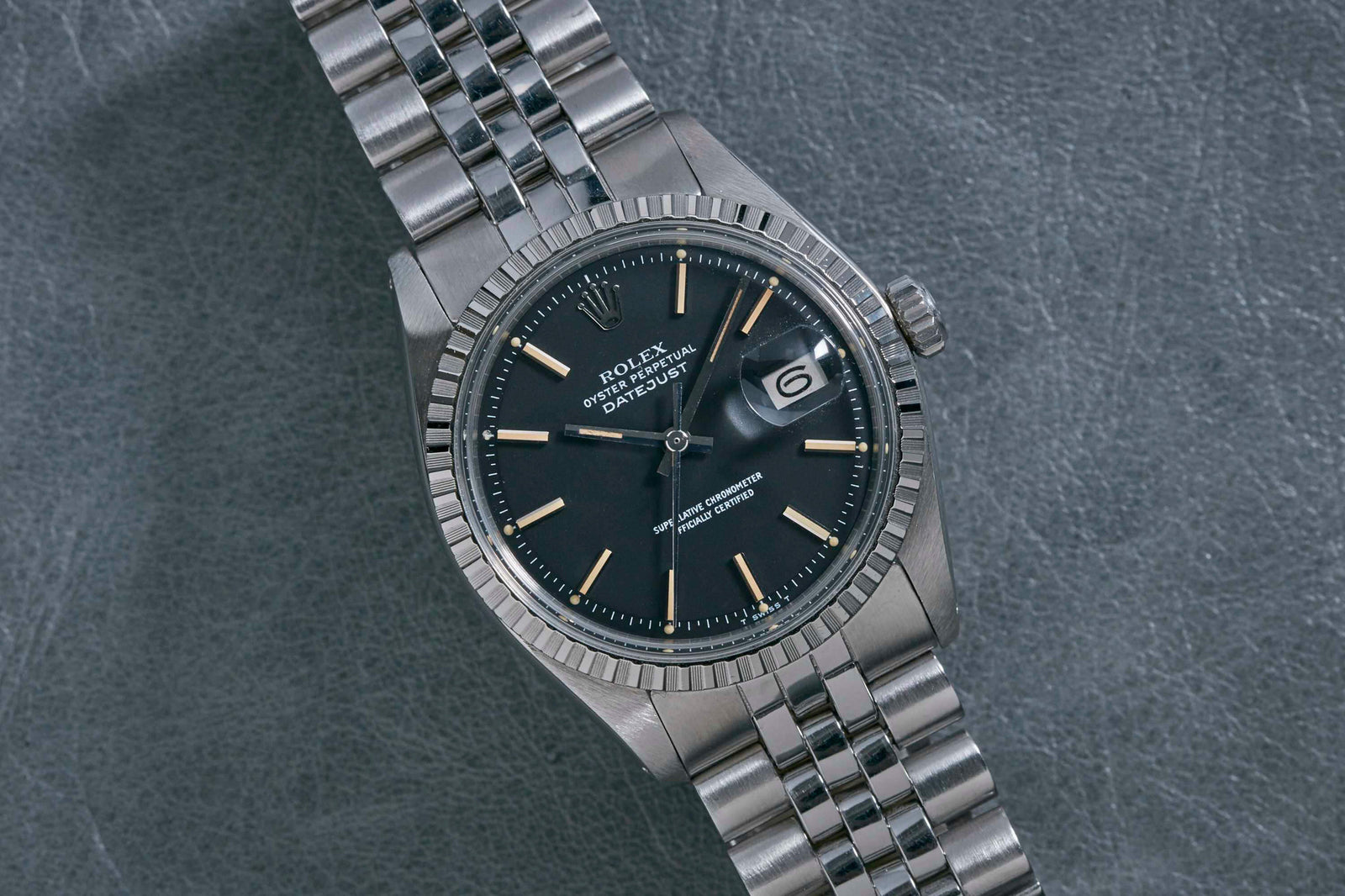 The Rolex Datejust by Rolex features a stainless steel case, black dial, date window at 3 o’clock, fluted bezel, and jubilee bracelet, elegantly displayed on a textured grey surface.
