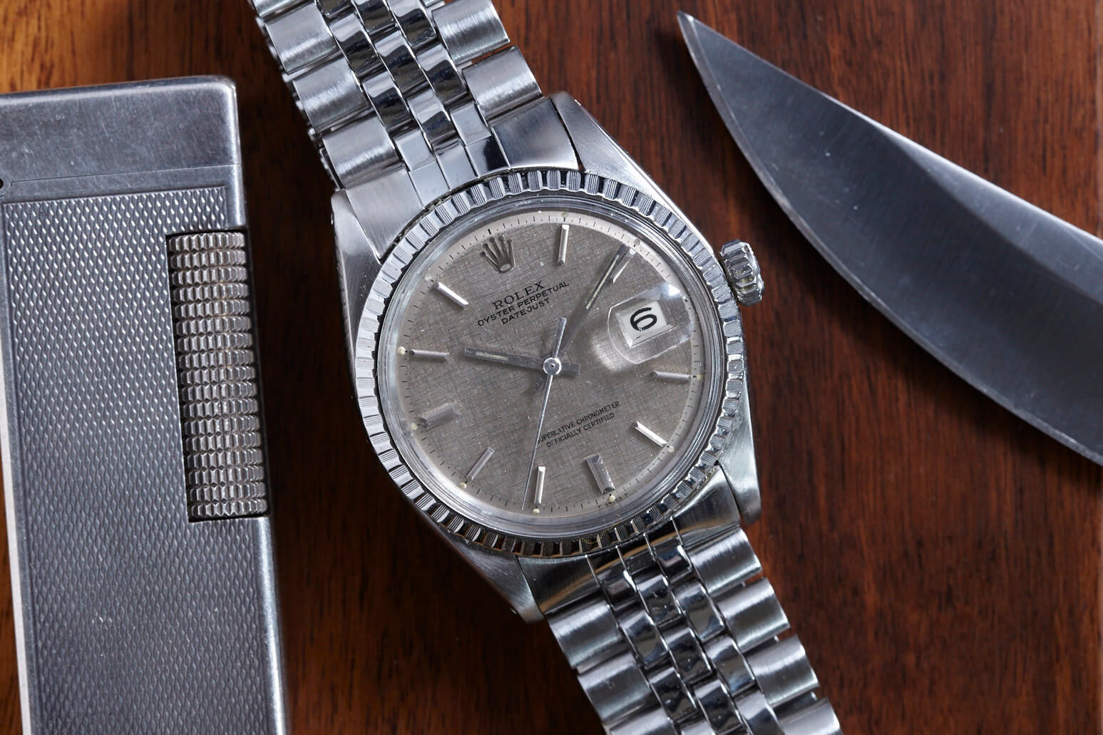 A Rolex Datejust 1601 Grey Linen wristwatch with a metal bracelet rests on a wooden surface beside a metal lighter and a shiny-bladed knife. The watch features automatic movement, date display, and a textured grey linen dial.