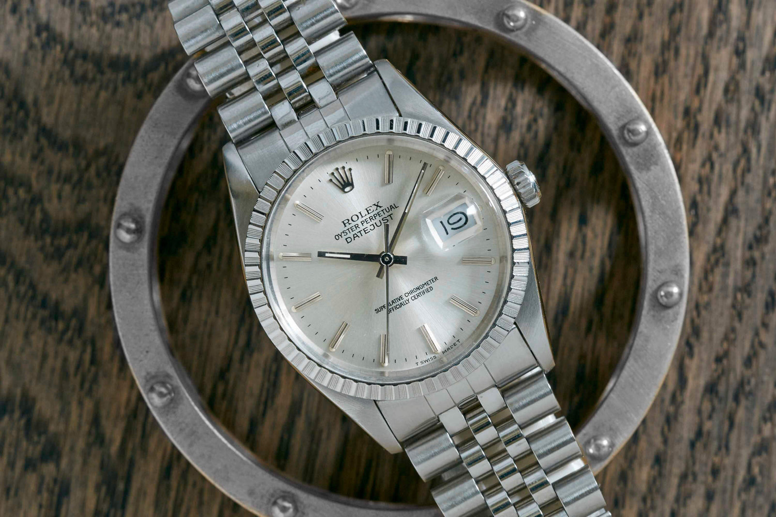 A silver Rolex Datejust by Rolex, featuring a jubilee bracelet, fluted bezel, and date display, rests on a wooden surface framed by a metal ring.