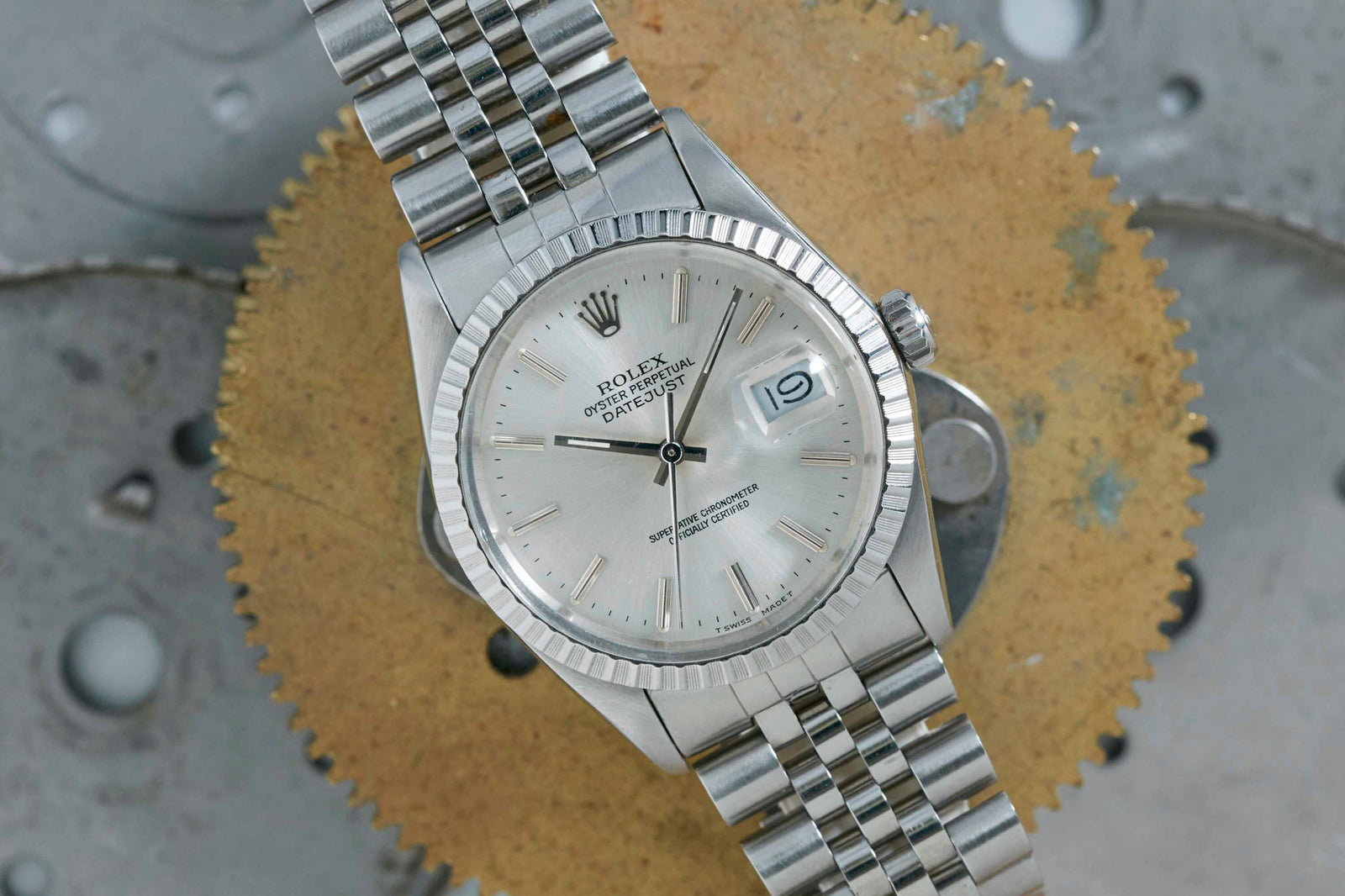 A silver Rolex Datejust by Rolex, featuring a fluted bezel and jubilee bracelet, sits atop a large, worn brass gear with a metallic backdrop. The iconic watch shows the date as the 19th.