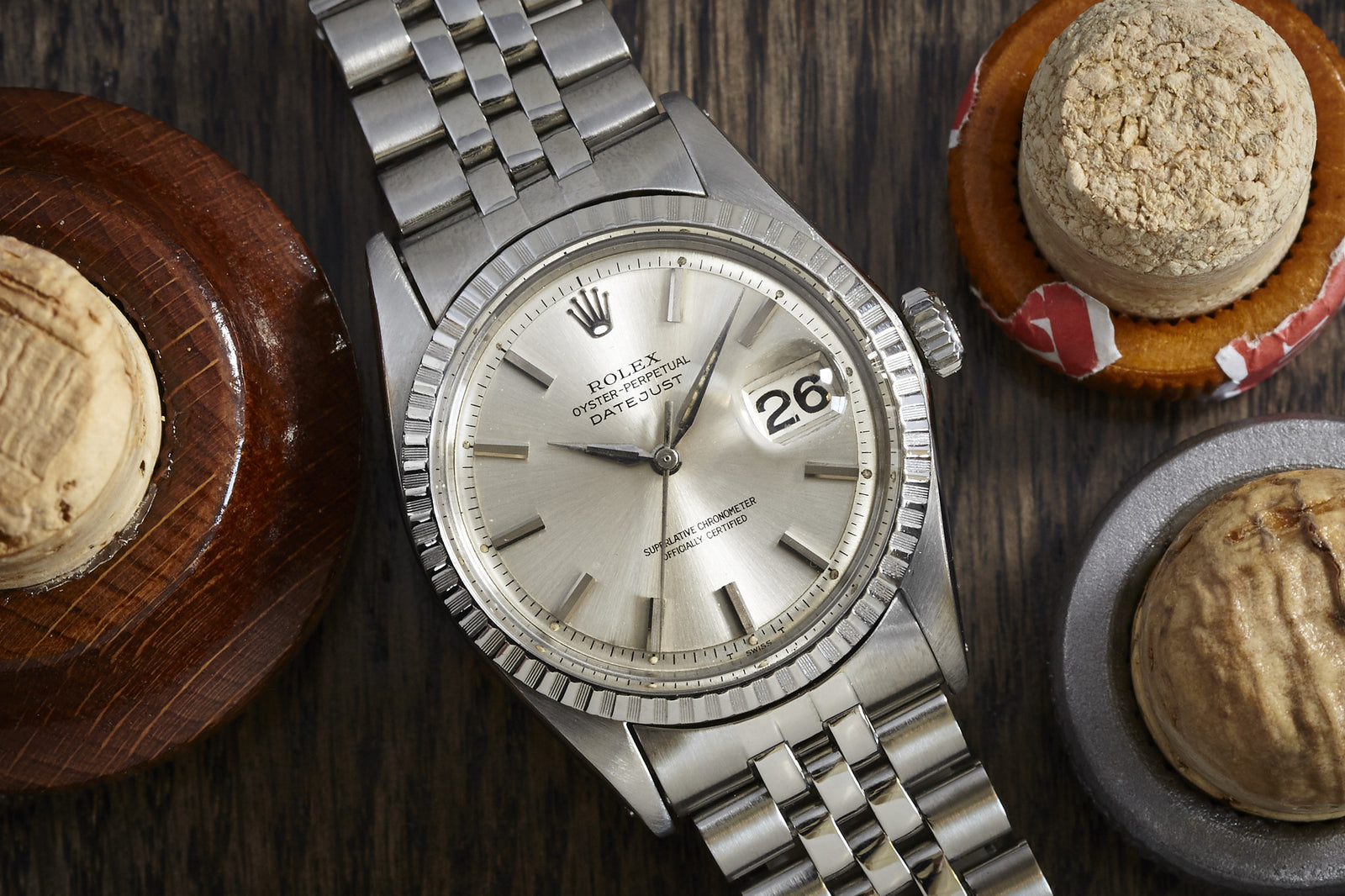 A Rolex Datejust by Rolex with Alpha hands and a stainless steel bracelet sits on wood, surrounded by three champagne corks.